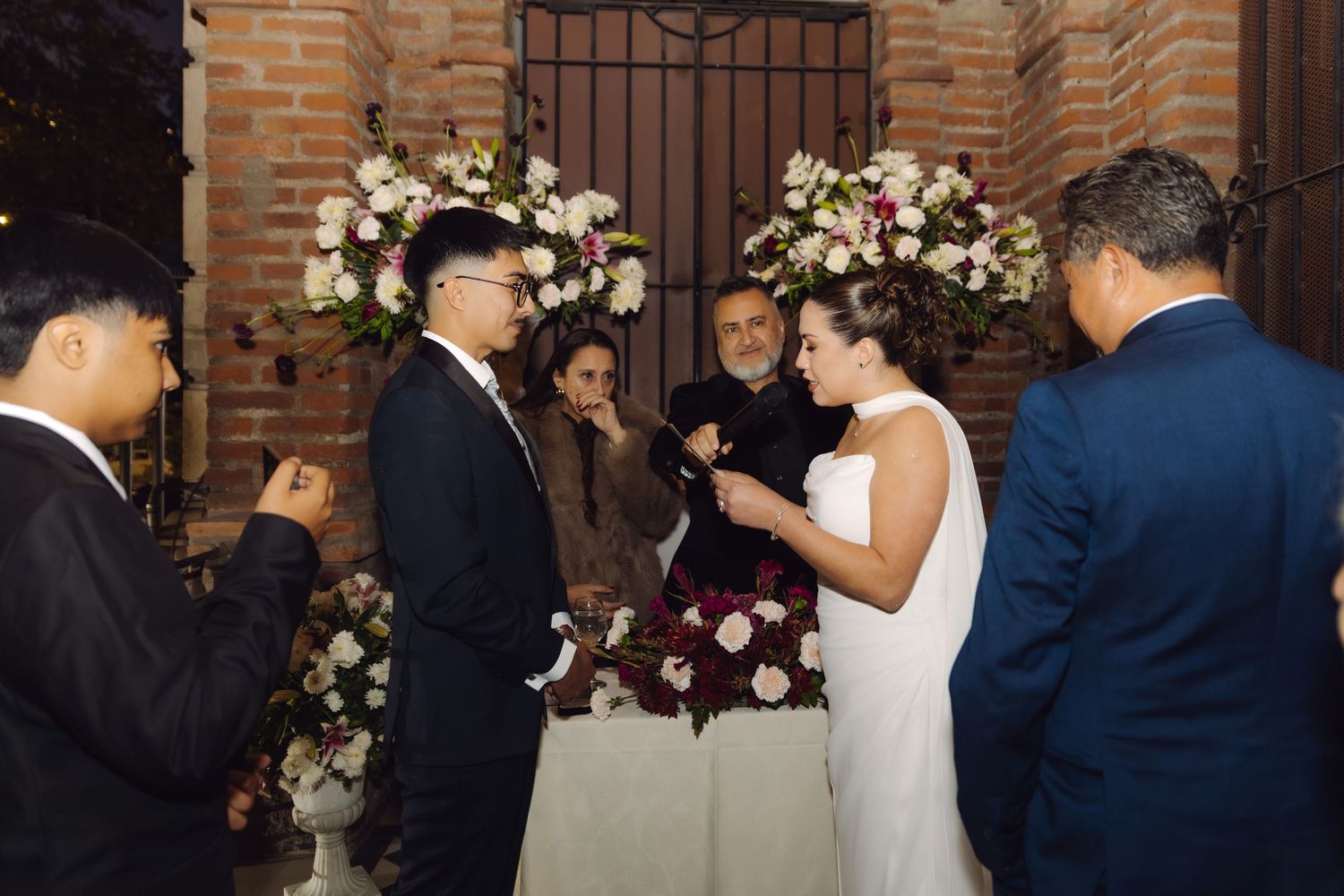 Bride and groom holding hands during wedding ceremony surrounded by guests and lush floral displays.