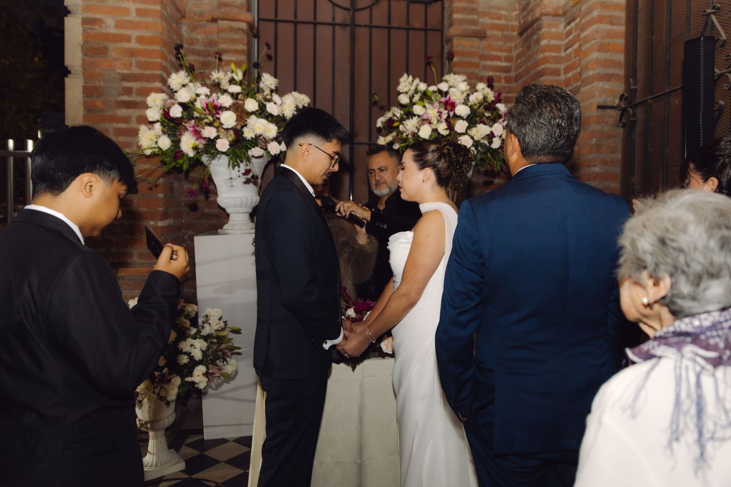 Groom placing ring on brides finger during wedding ceremony with brick wall and floral backdrop.