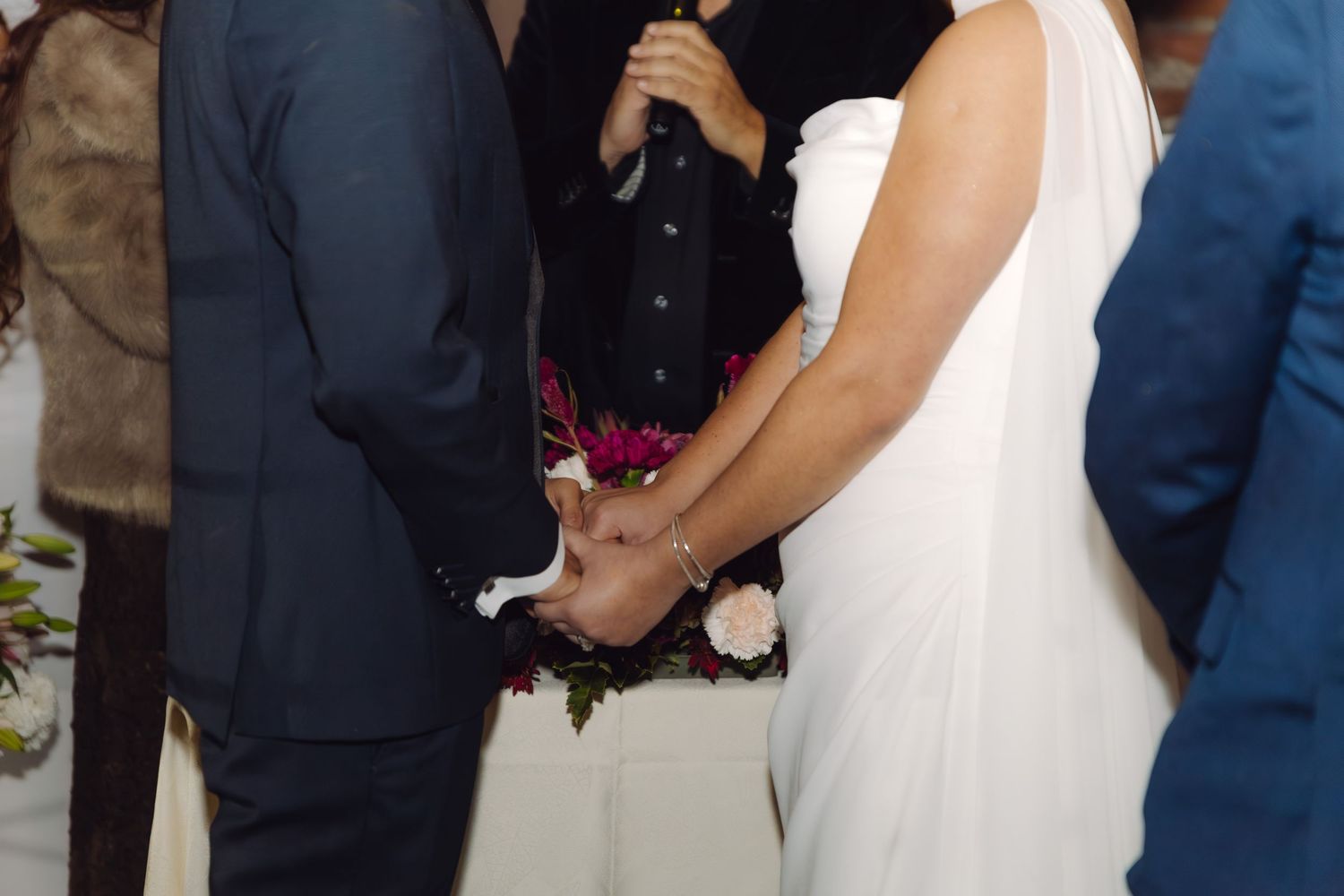 Close-up of bride and groom holding hands during wedding ceremony in white and burgundy decor.
