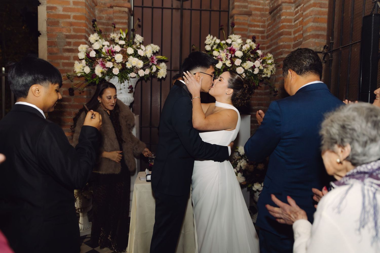 Bride and groom share first kiss at wedding ceremony in front of brick wall with flowers.