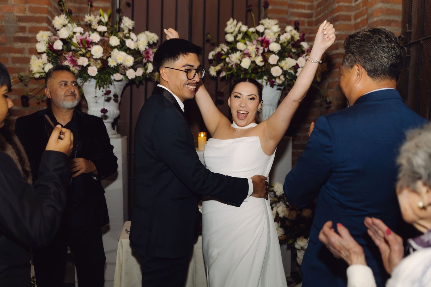 Joyful bride raising arms in celebration with groom after wedding ceremony with floral backdrop.