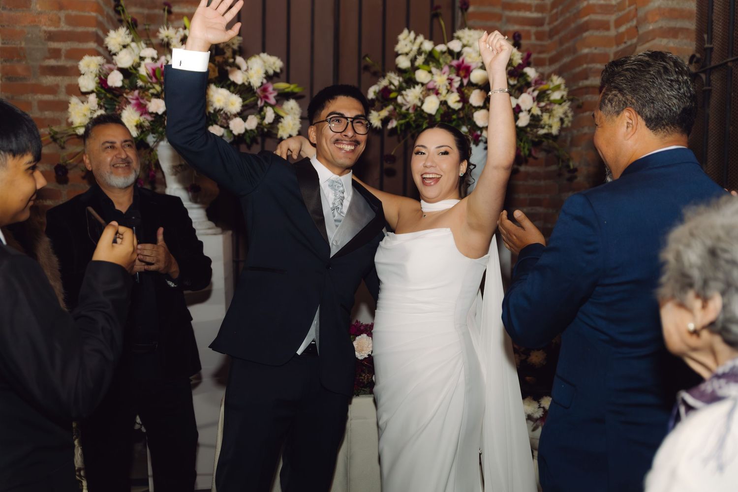 Joyful newlyweds raise hands in celebration walking down aisle in brick church surrounded by clapping guests.