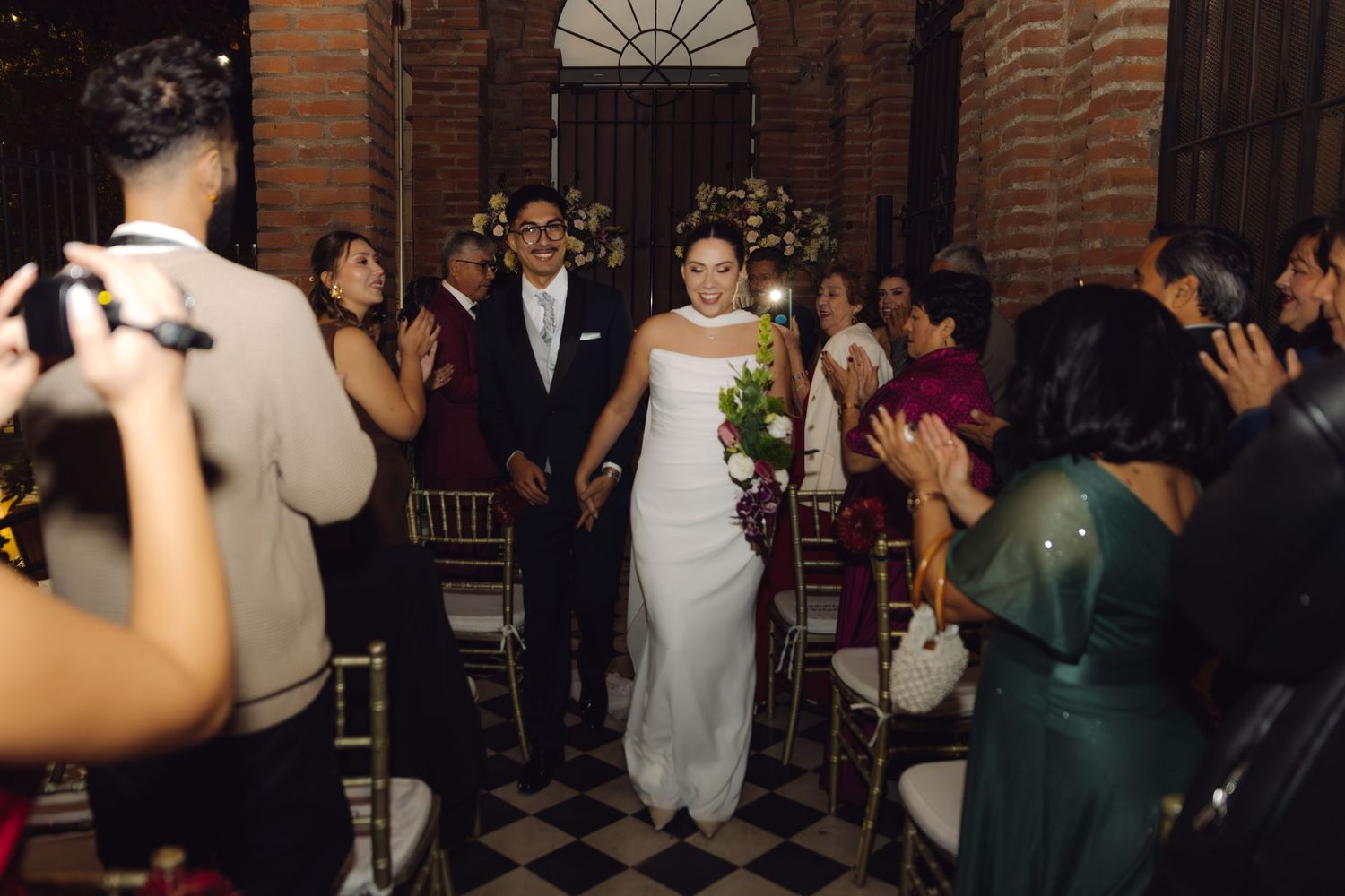 Bride and groom walk hand in hand down checkered aisle while wedding guests applaud in elegant venue.