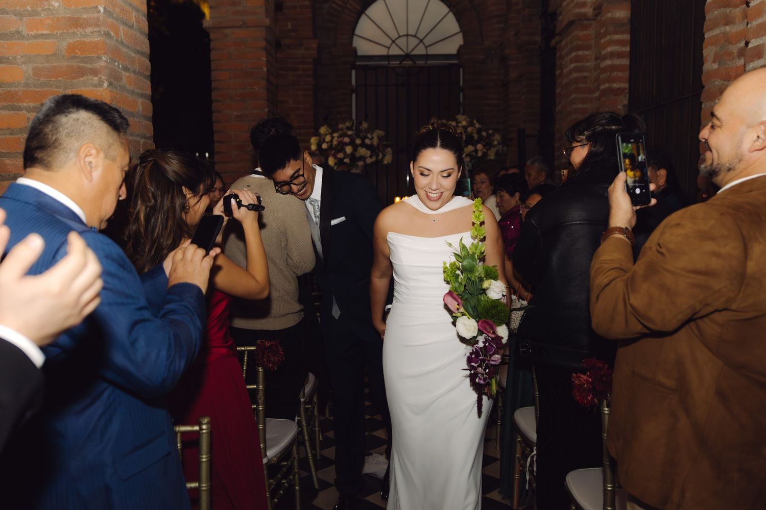 Bride in white gown looks down holding bouquet while walking through ceremony aisle lined with guests.