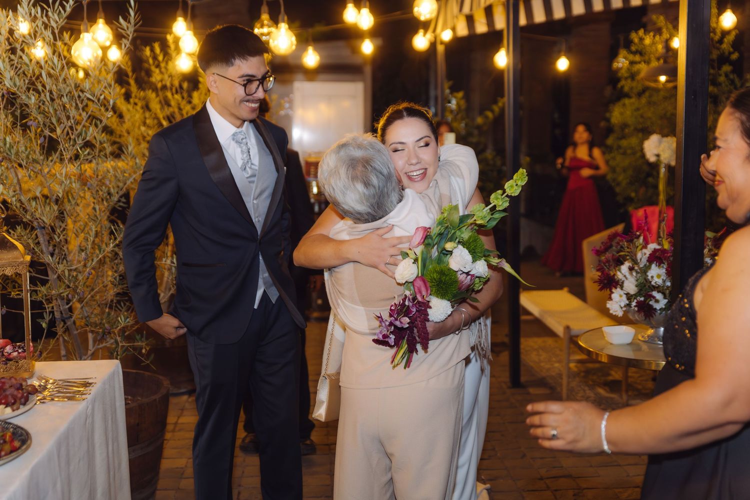 Bride smiles while receiving warm hug from elderly guest outdoors at evening wedding reception with lights.