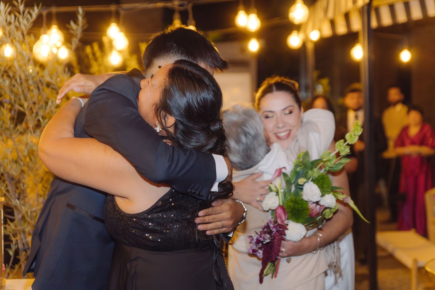 Groom embraces family member while smiling bride holds bouquet at warmly lit outdoor wedding reception.
