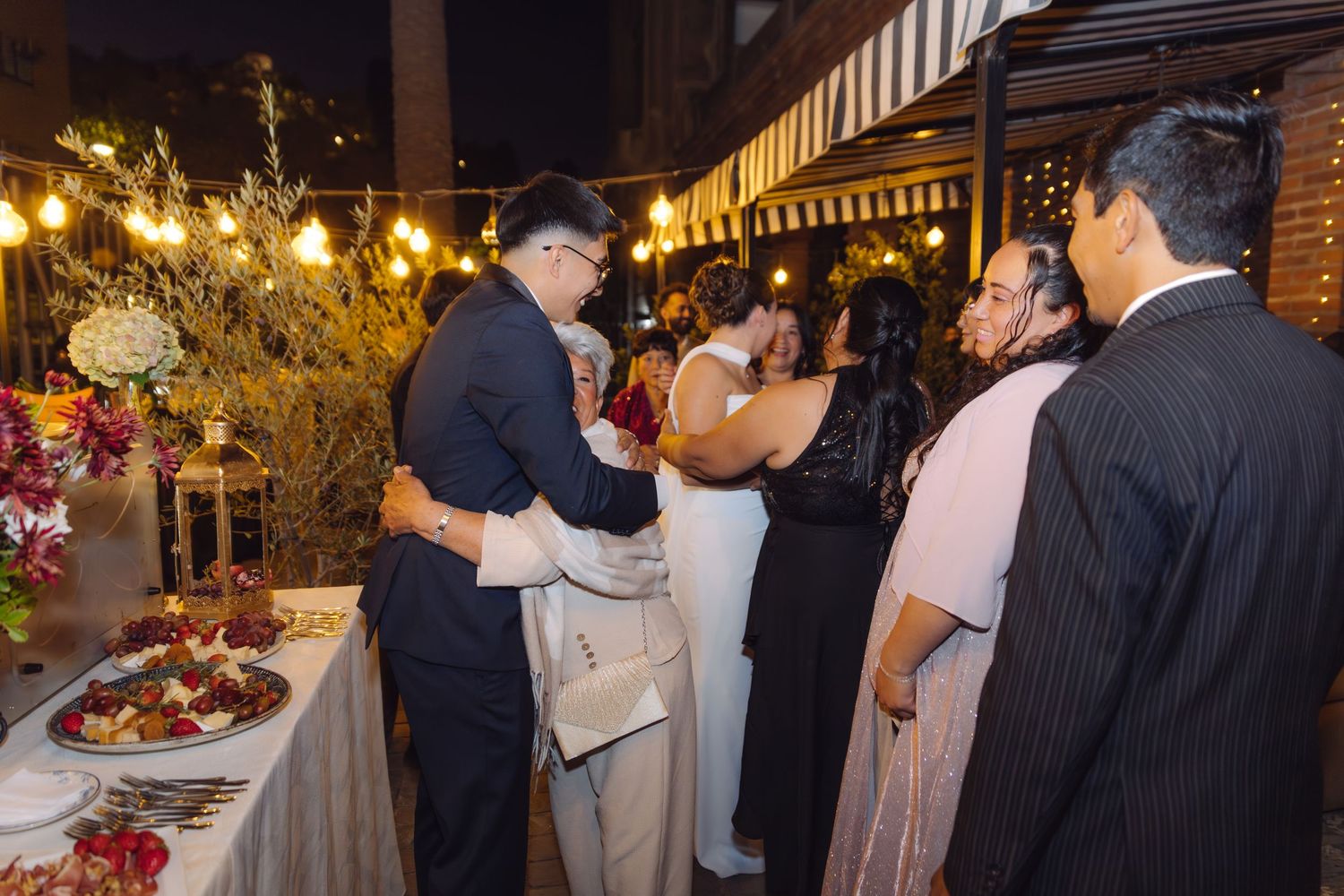 Newlyweds greet guests at outdoor evening wedding reception under string lights near decorated table.
