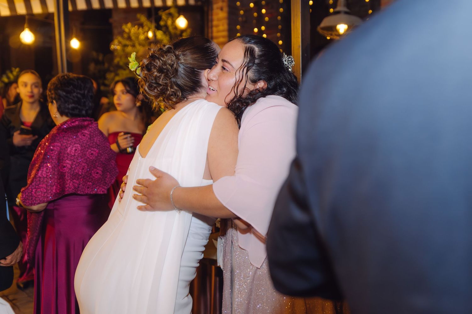 Bride in white gown embracing a guest in a pink dress at a warmly lit wedding reception.