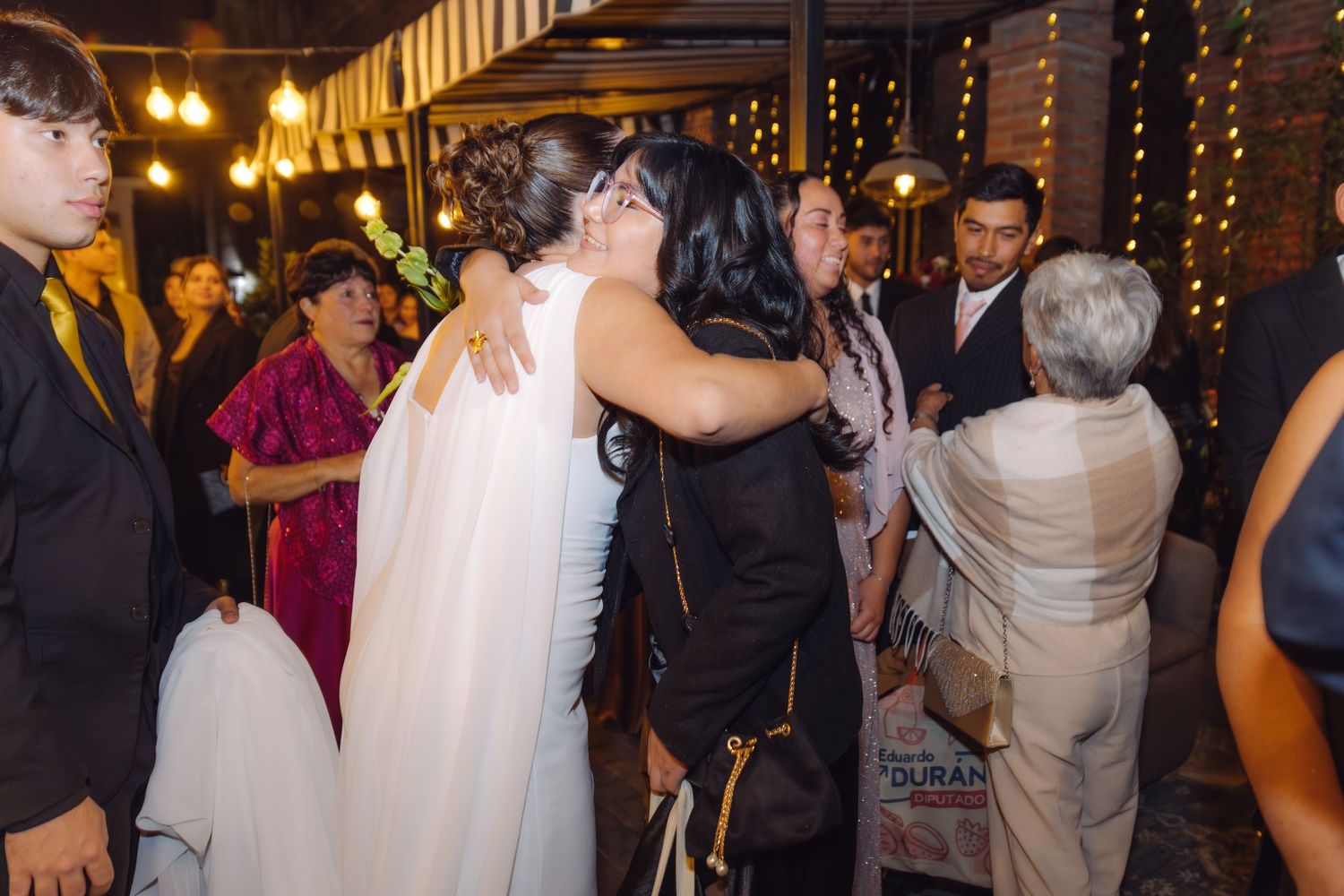Bride in white wedding gown hugging a guest in black at an outdoor evening wedding reception.