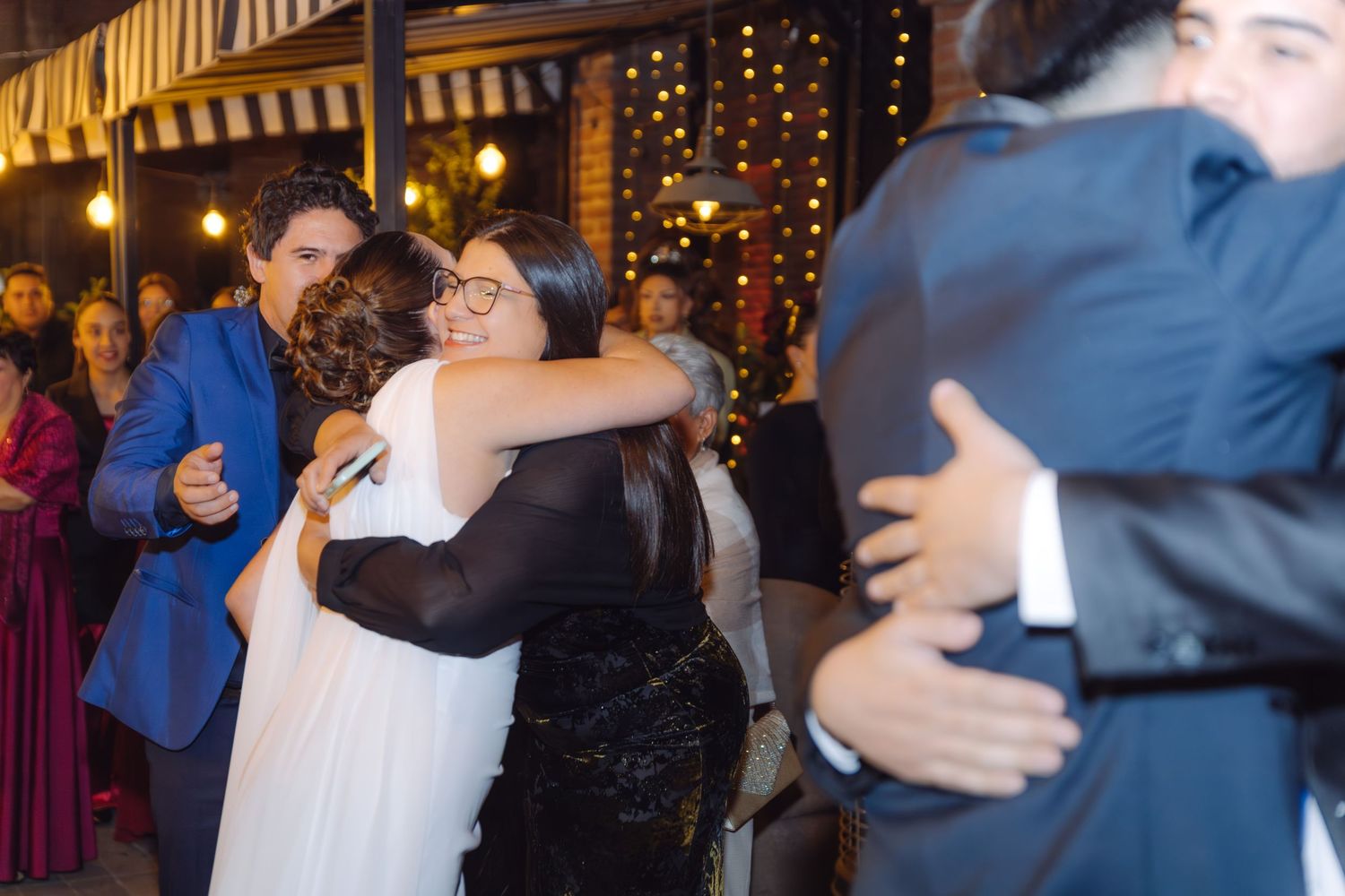 Bride in white dress sharing a warm hug with a guest in black at a festive wedding celebration.