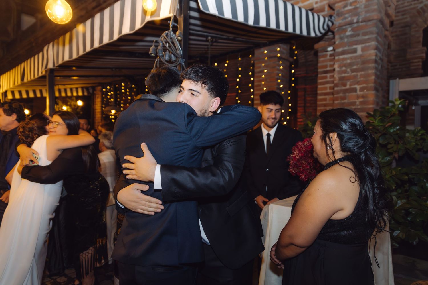Two men in suits sharing an emotional embrace at an outdoor evening wedding reception.