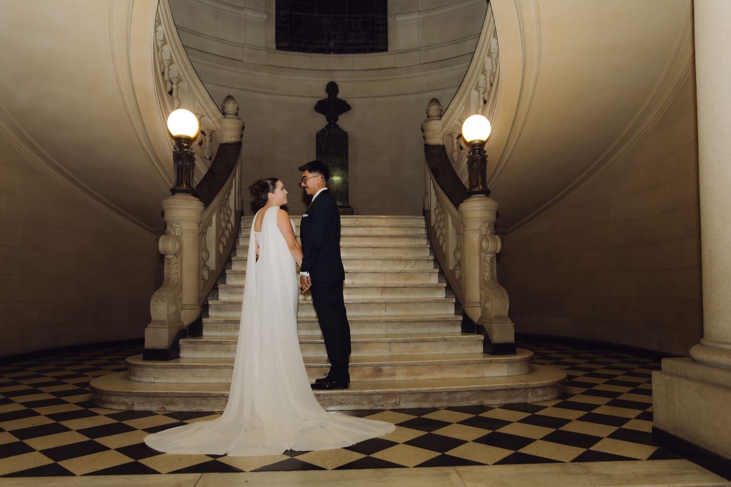 Bride and groom standing face to face at the base of an elegant marble staircase at night.