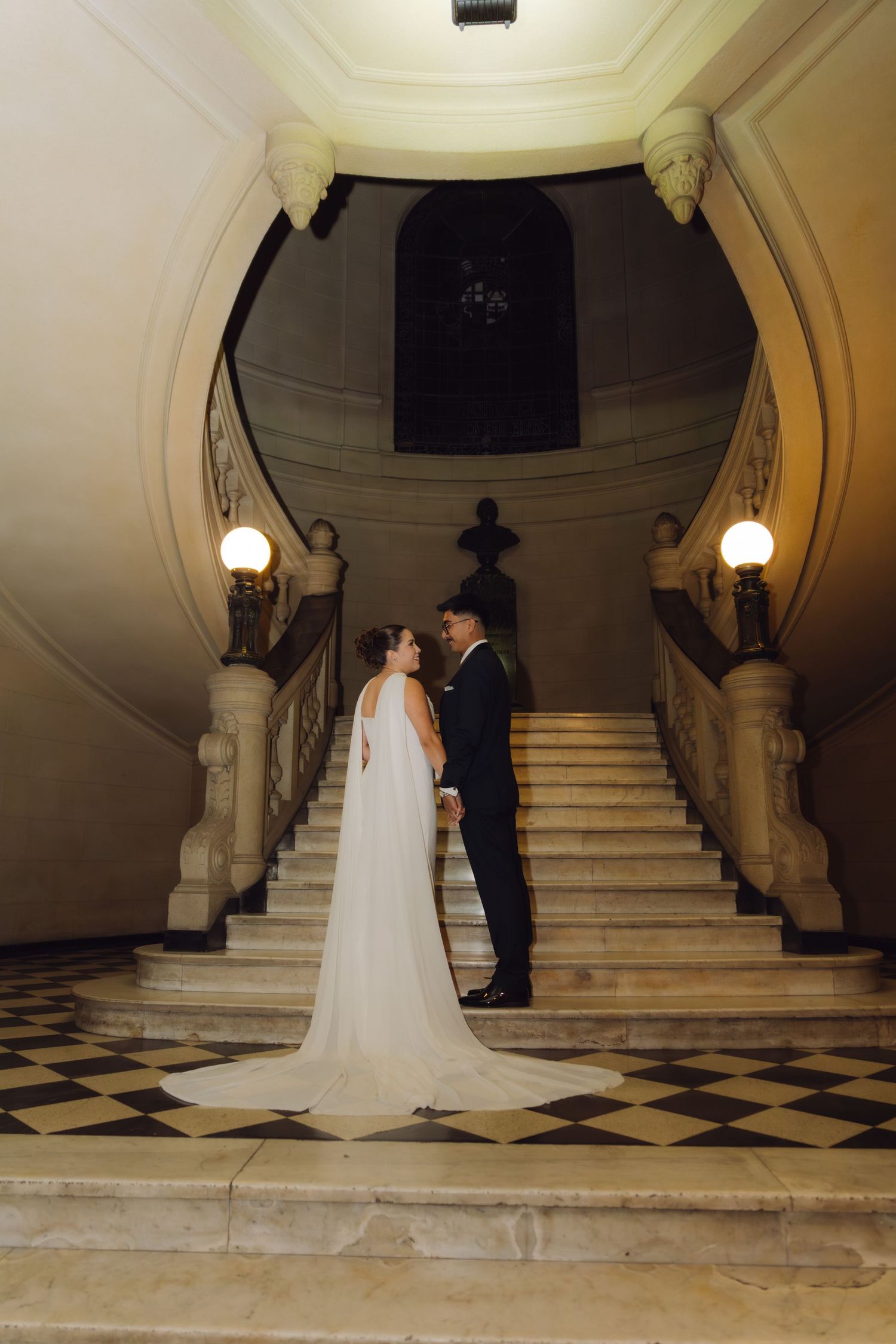 Bride in white gown with long train and groom in black suit posing on grand marble staircase.