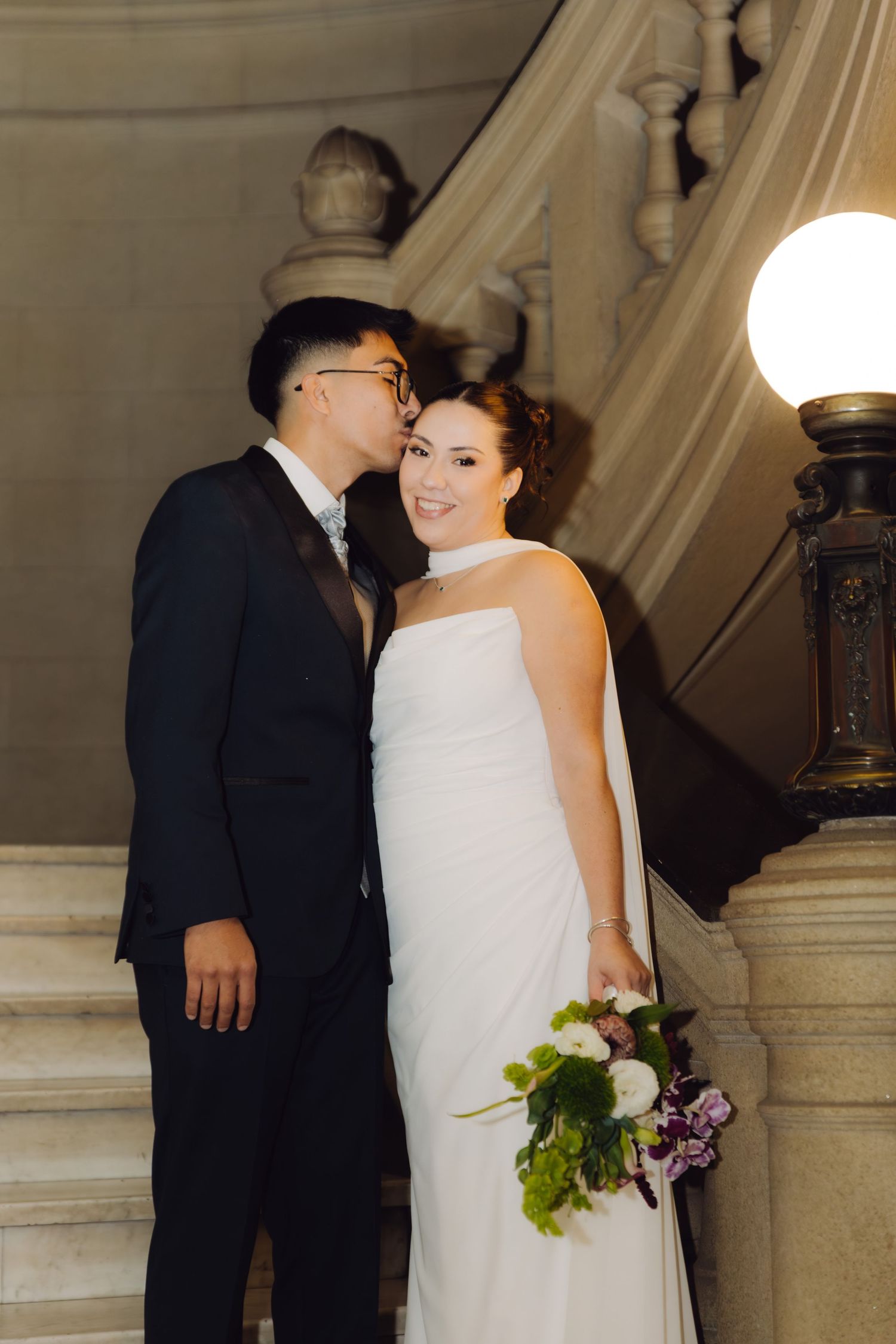 Groom in black suit kissing bride on cheek as she holds a lush floral bouquet on marble stairs.