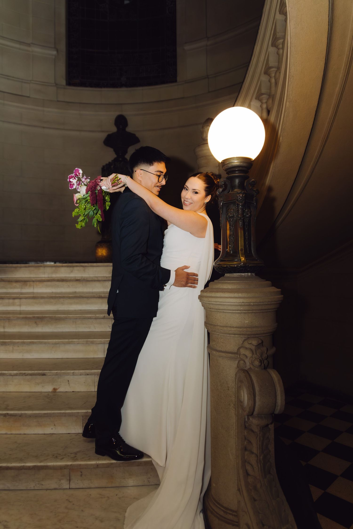 Bride and groom embracing romantically on an elegant marble staircase with a glowing lamp.