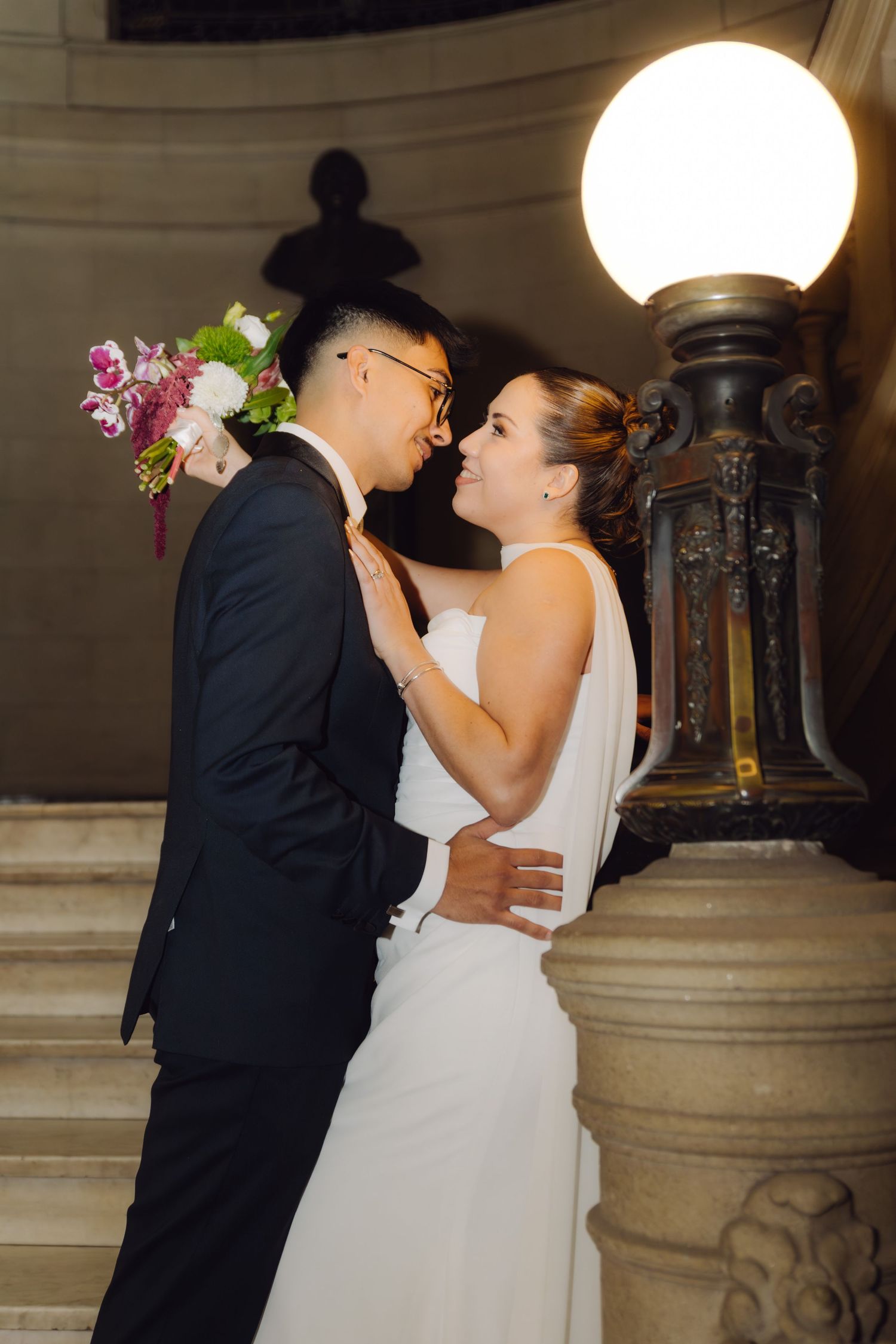 Bride and groom gazing at each other lovingly beside a glowing lamp on a grand marble staircase.