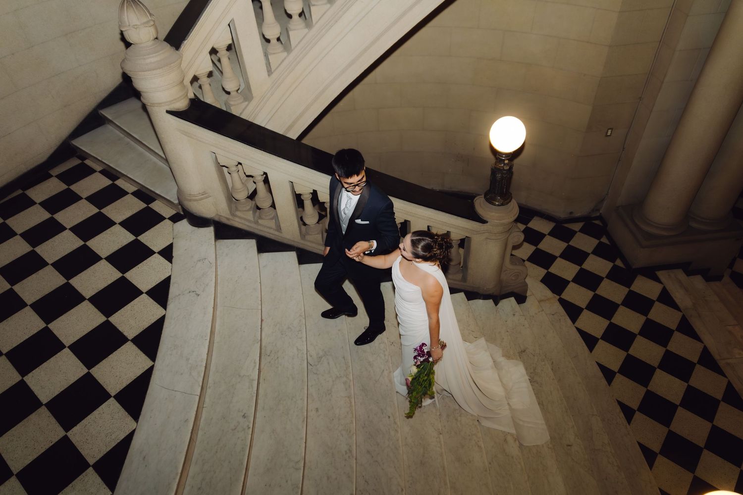 Bride and groom walking down grand marble staircase with checkered floor at elegant wedding venue.