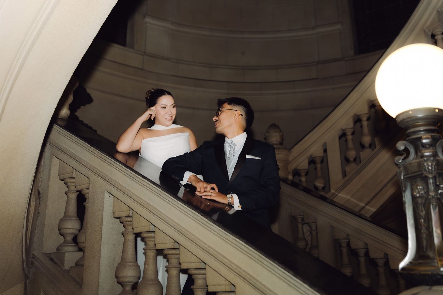 Bride and groom sharing romantic moment on ornate marble staircase balcony at night wedding.