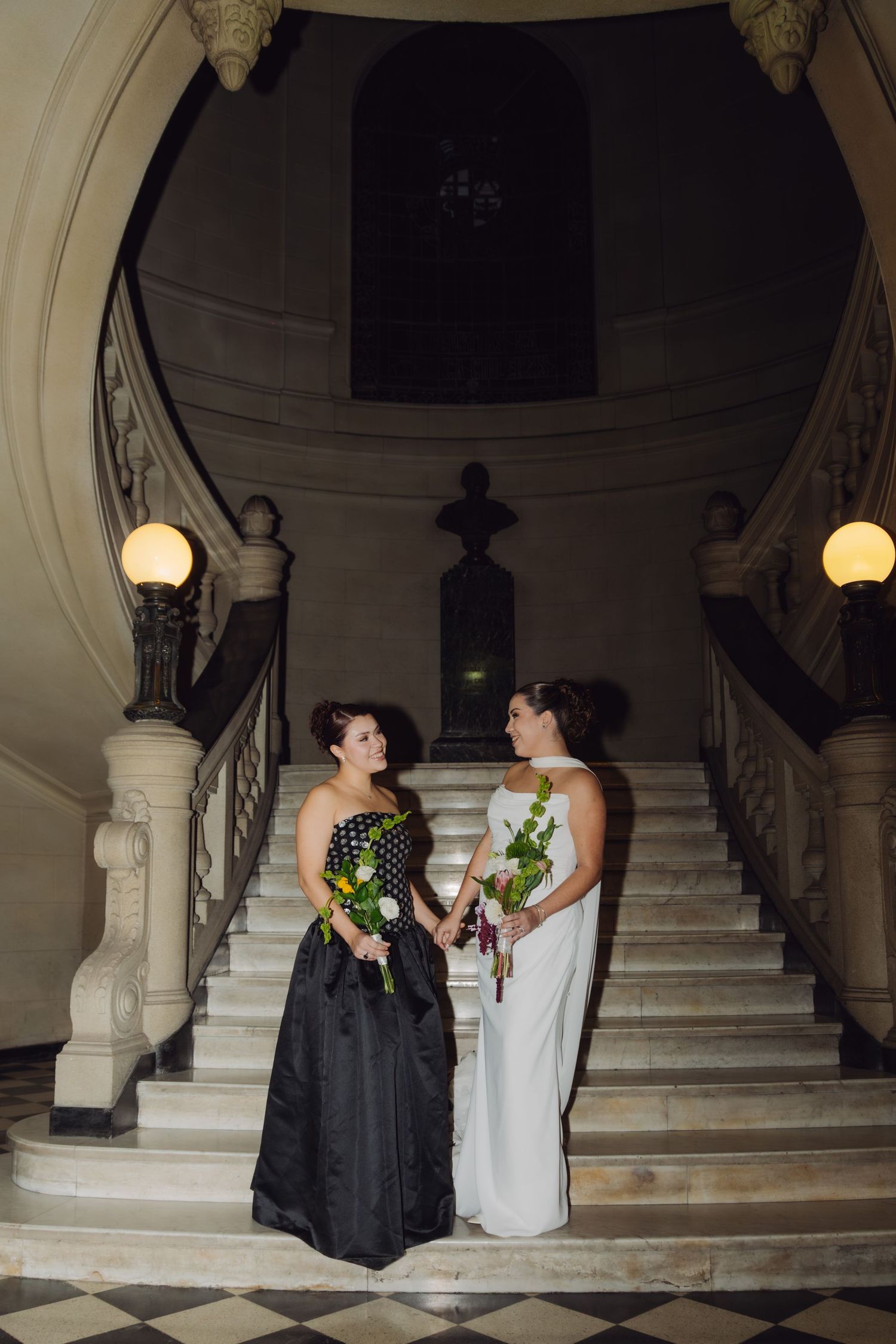 Two women in black and white gowns holding bouquets on ornate marble wedding staircase.
