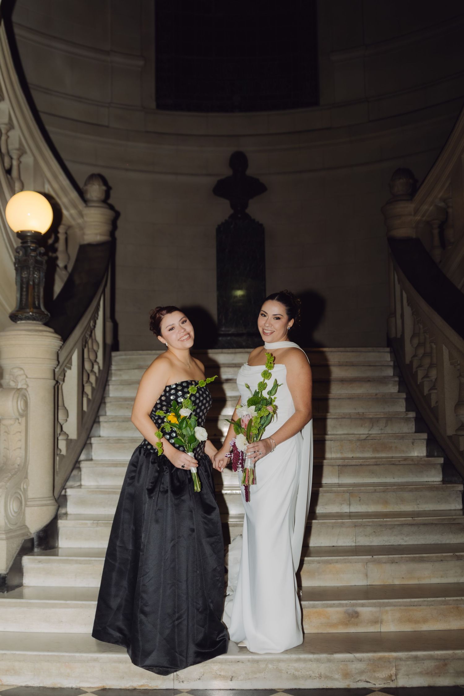 Bridesmaid in black gown and bride in white dress posing with bouquets on marble stairs.