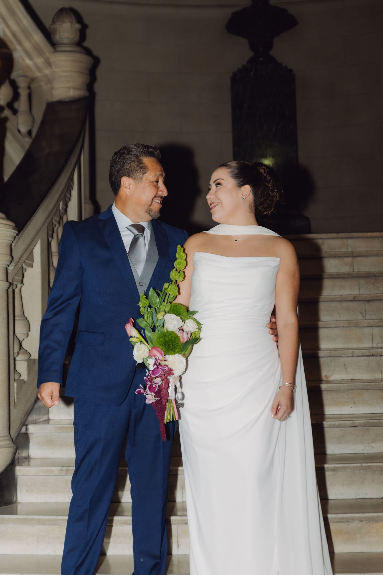 Bride and groom sharing loving glance while holding bouquet on elegant marble staircase.