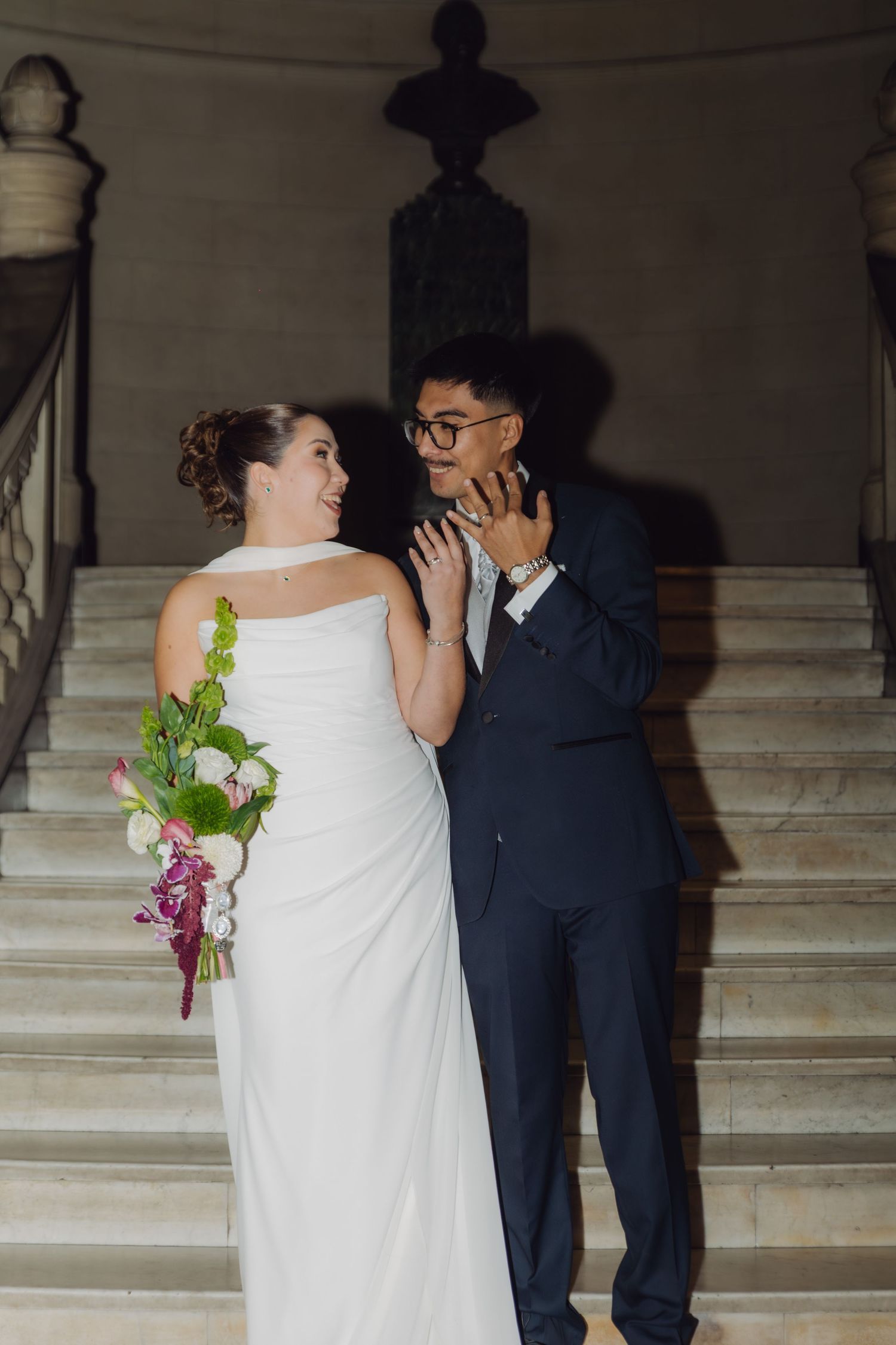 Bride tenderly touching grooms face while holding bouquet on grand marble wedding staircase.