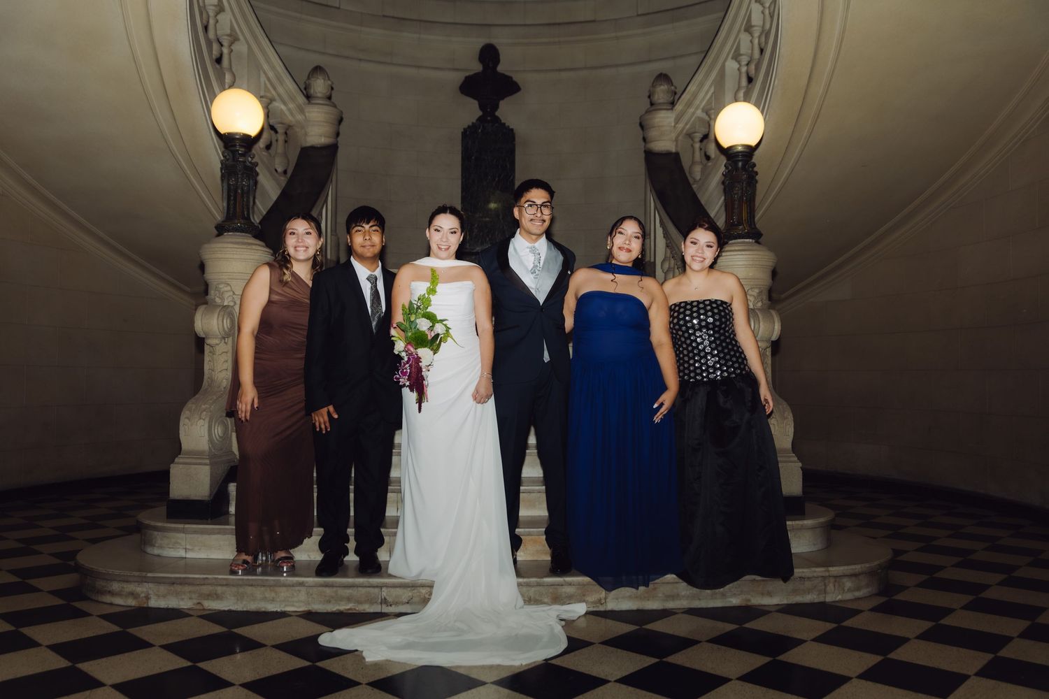 Wedding party group portrait on checkered marble floor beneath ornate classical staircase.