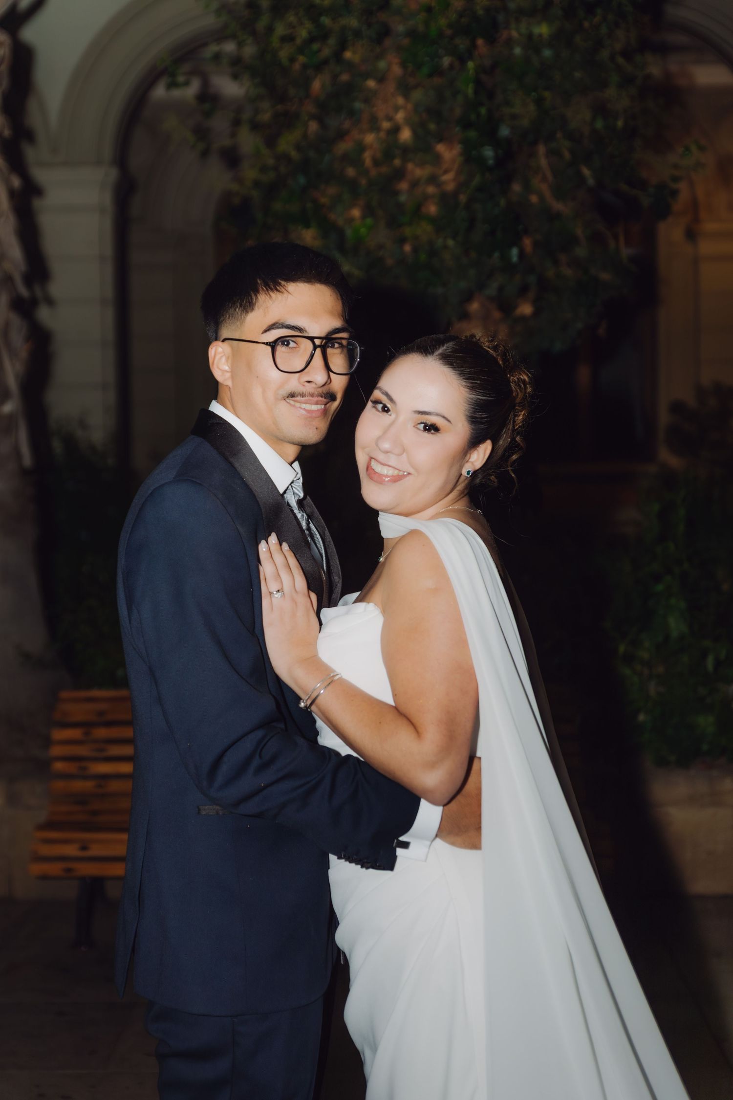 Smiling bride in white gown with long veil poses with groom in navy suit for wedding portrait at night.