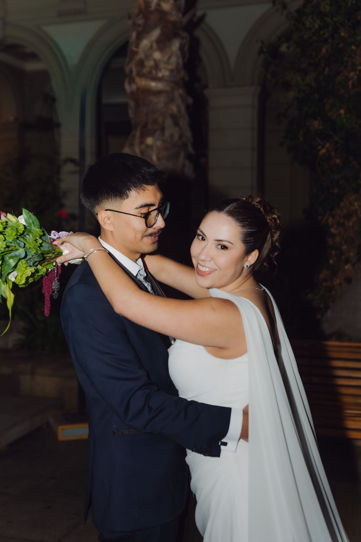 Bride in white sleeveless gown embraces groom holding bouquet during nighttime outdoor wedding photo session.
