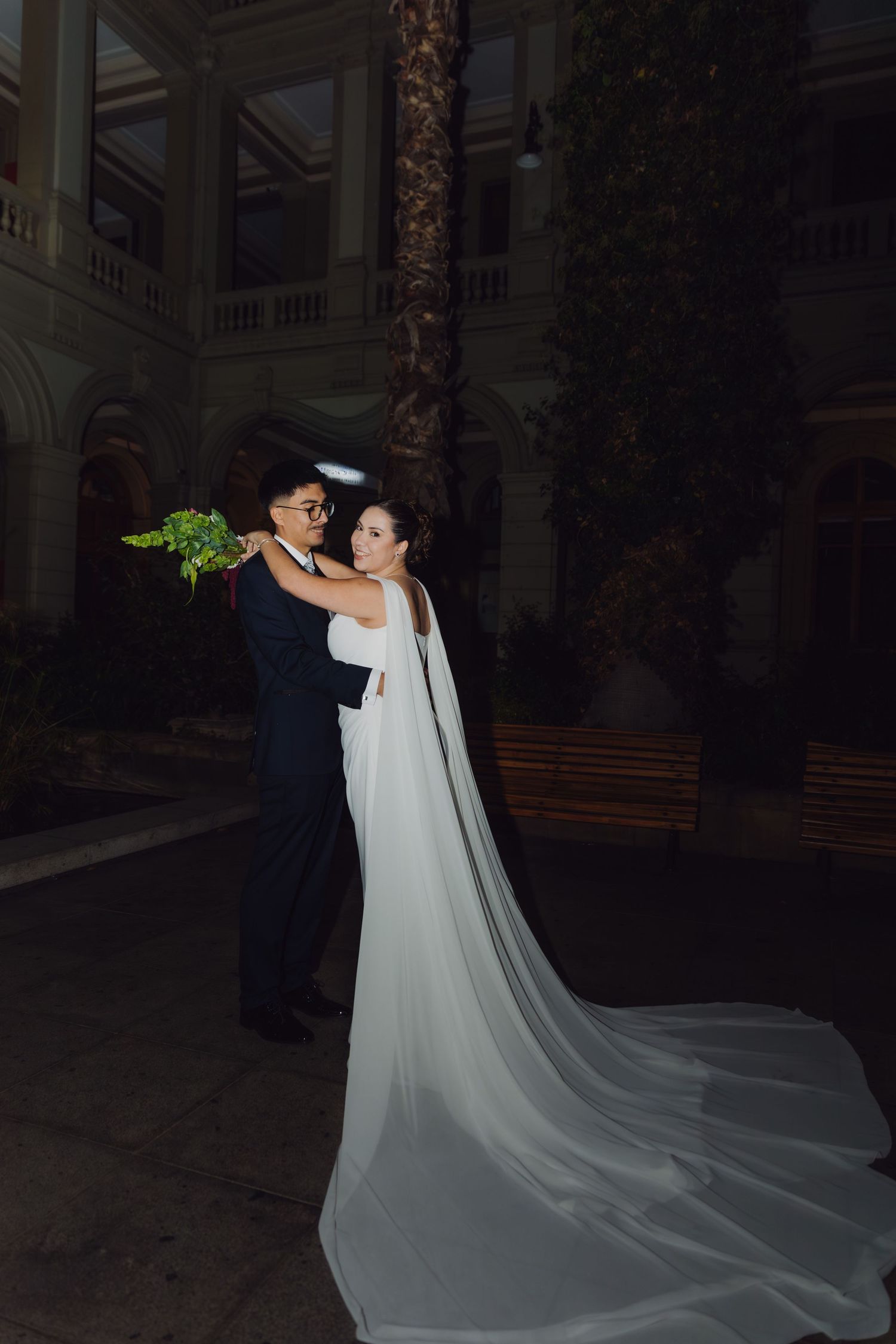 Bride in stunning white gown with flowing cathedral veil poses with groom outside elegant venue at night.