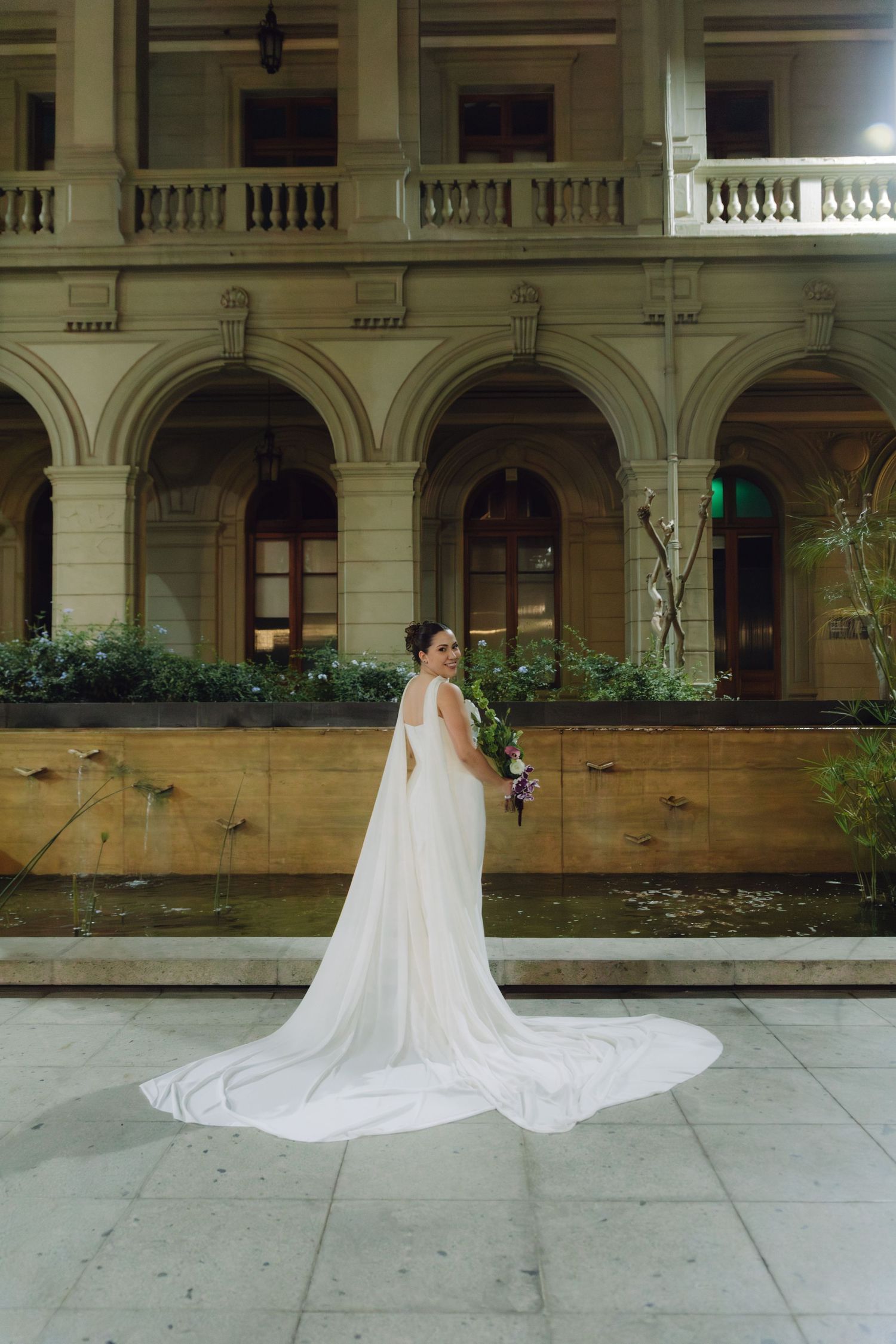 Elegant bride in white gown with dramatic cathedral veil poses solo outside a grand arched building at night.