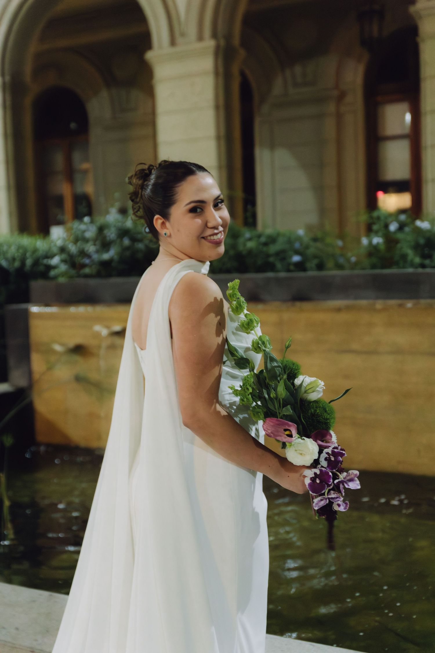 Smiling bride in white backless gown holds colorful bouquet of purple and white flowers at night venue.