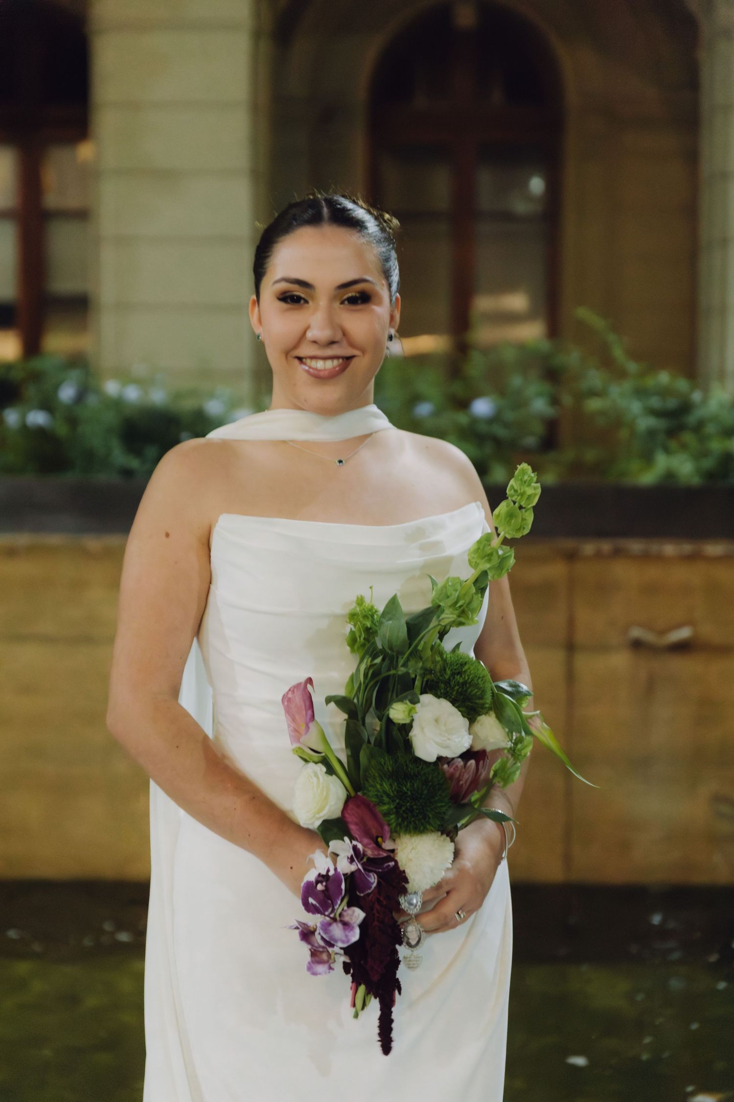 Radiant bride in white strapless gown smiles while holding lush bouquet of white, pink, and purple flowers.