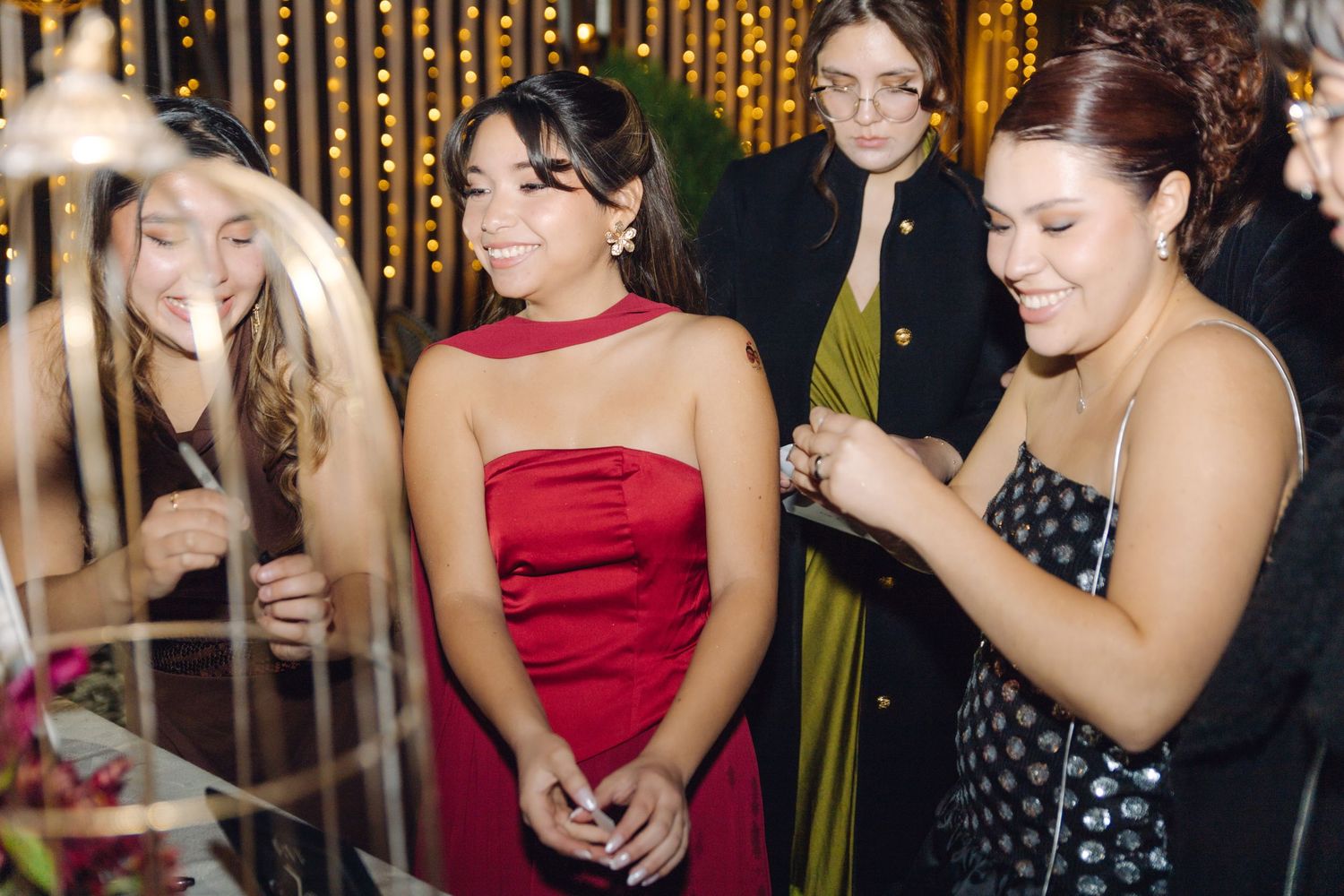 Young women in formal attire laughing and socializing at an elegant evening event with warm string lights.