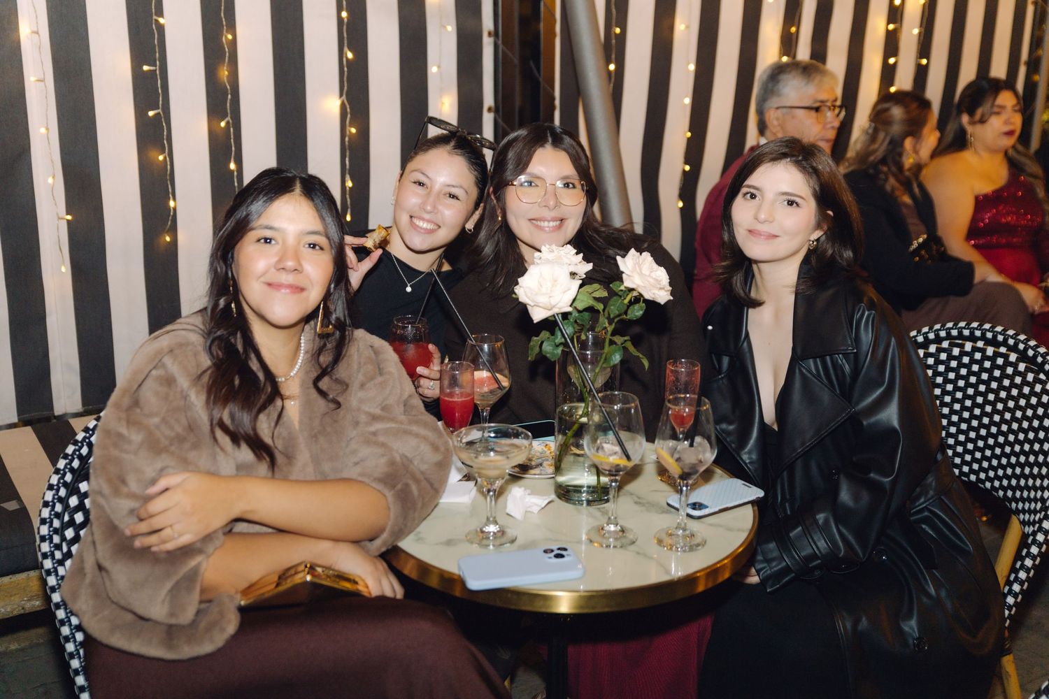 Four women smiling together at a table with cocktails and flowers at a stylish striped-wall restaurant.