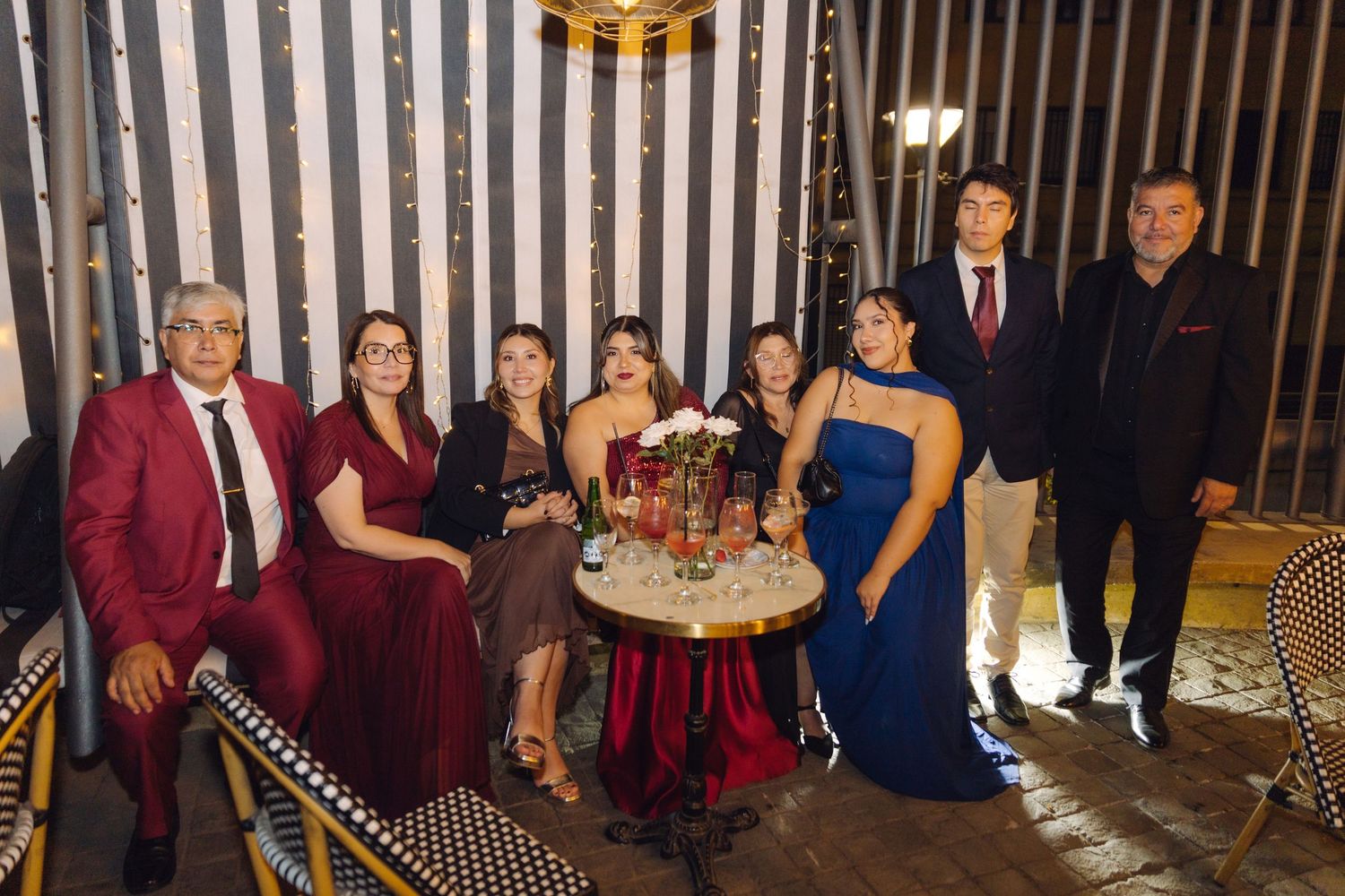 Group of eight adults in formal wear posing together at a festive evening celebration with string lights.