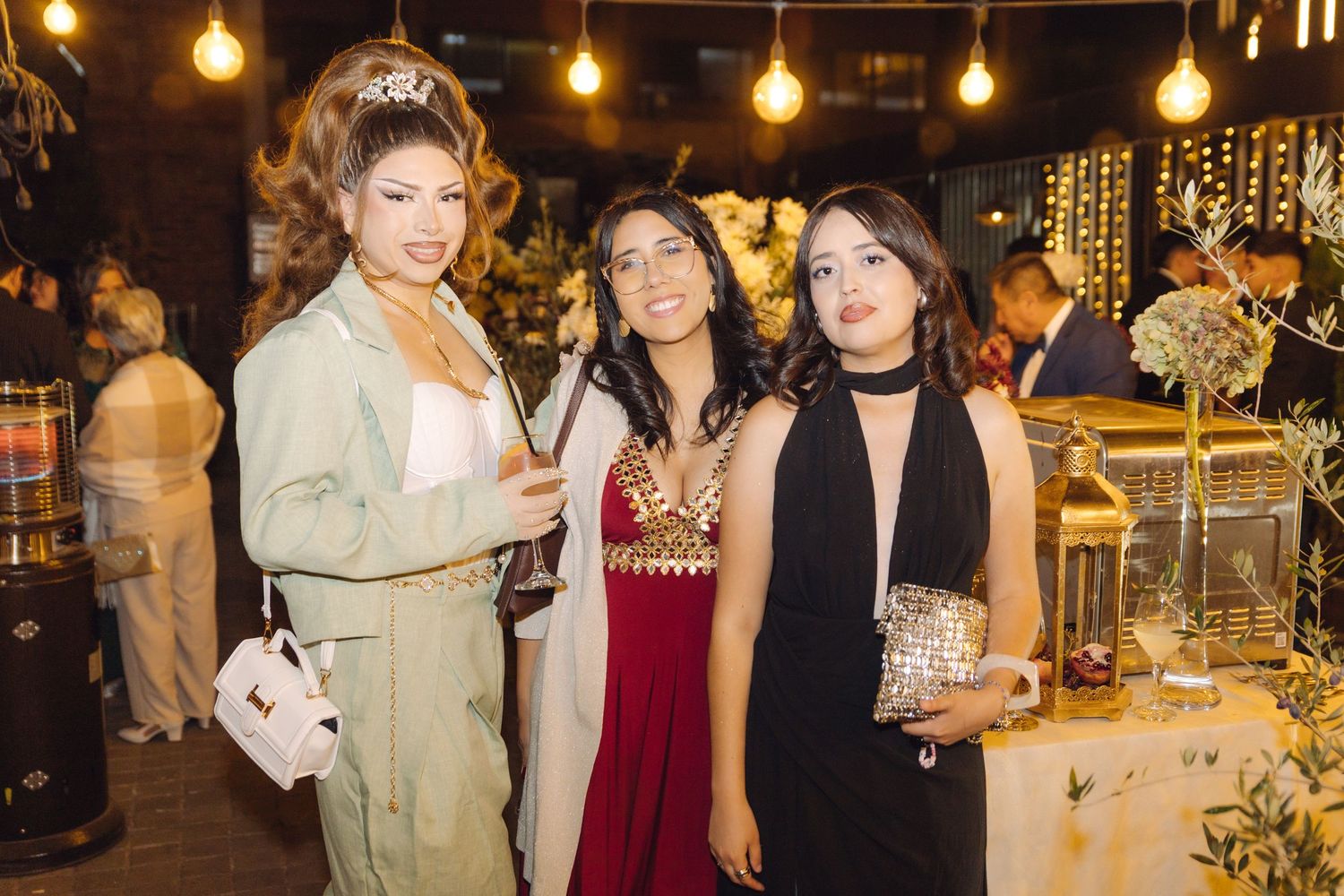 Three women in elegant formal outfits posing together at a glamorous evening party with warm lighting.