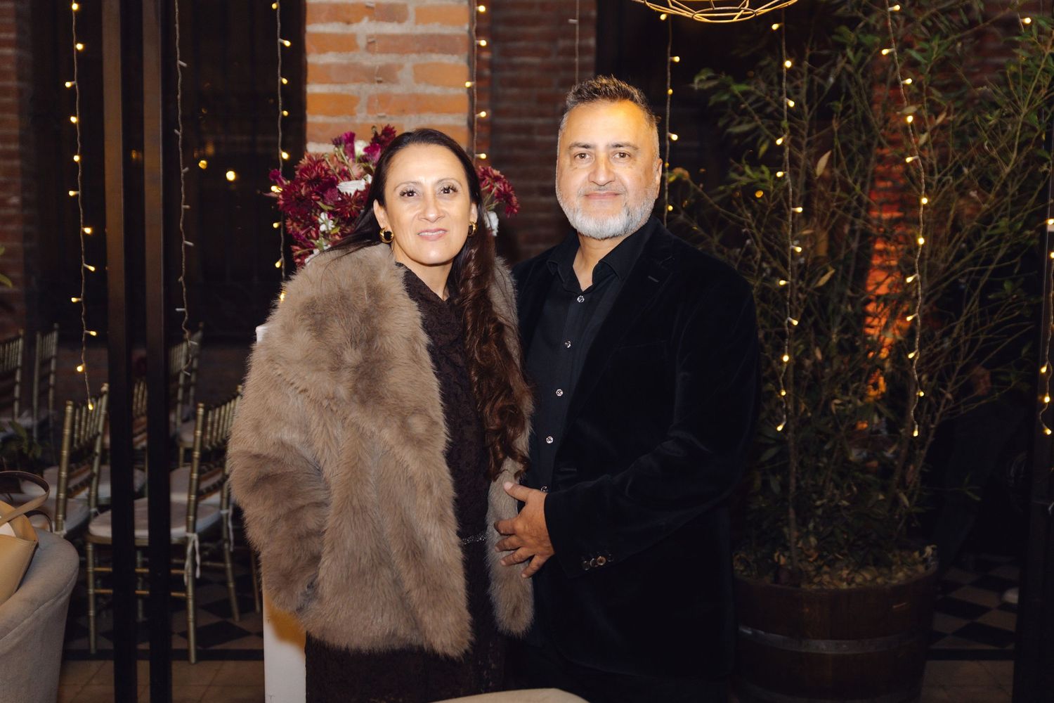 Elegant couple smiling together at an outdoor evening event surrounded by warm lights and greenery.