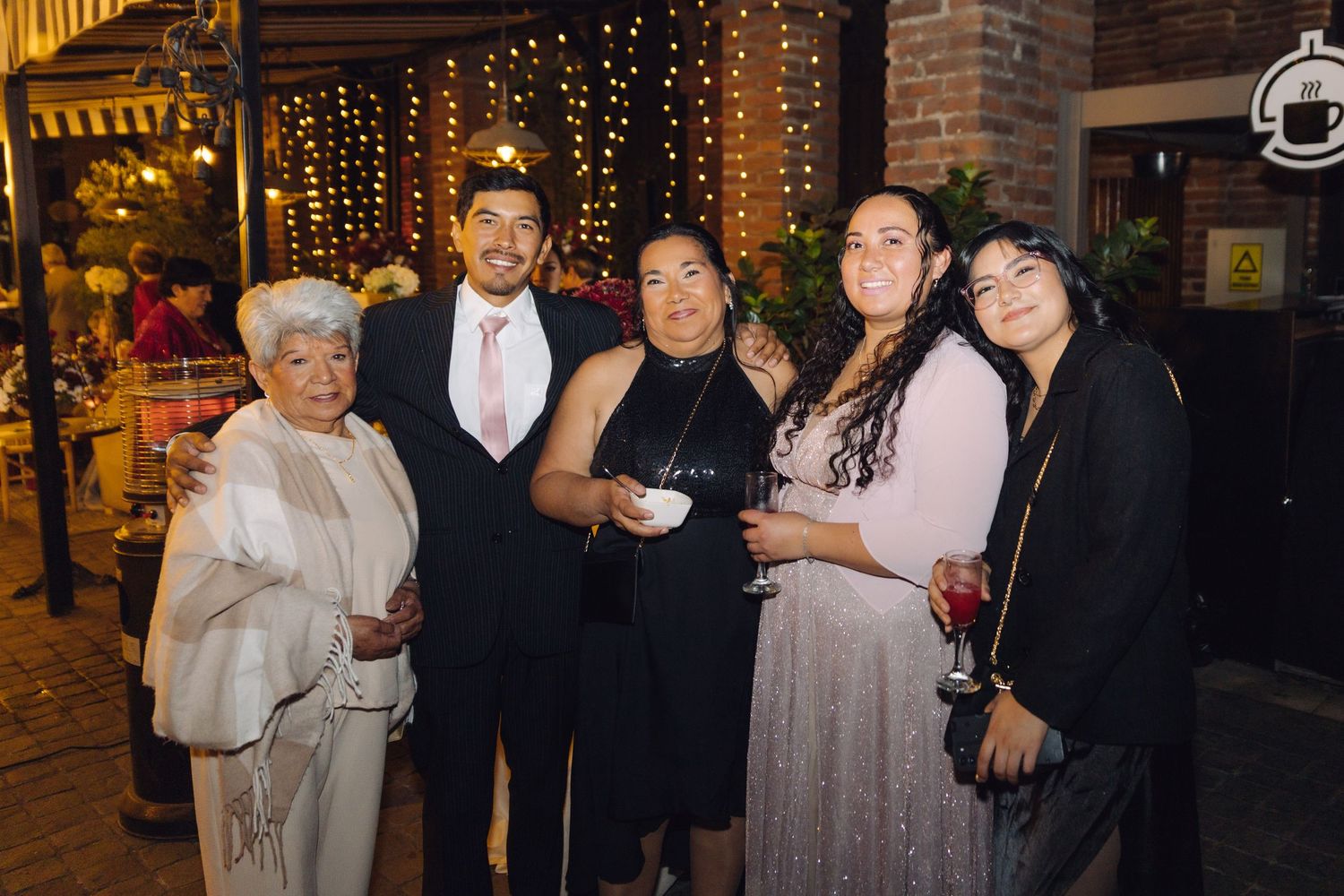 Group of six people in formal attire smiling together at an outdoor evening celebration with string lights.