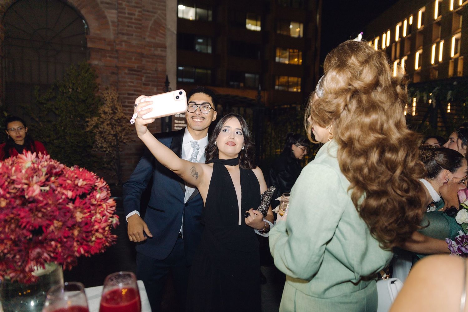 Man and woman taking a selfie at an elegant evening event with red floral centerpieces nearby.