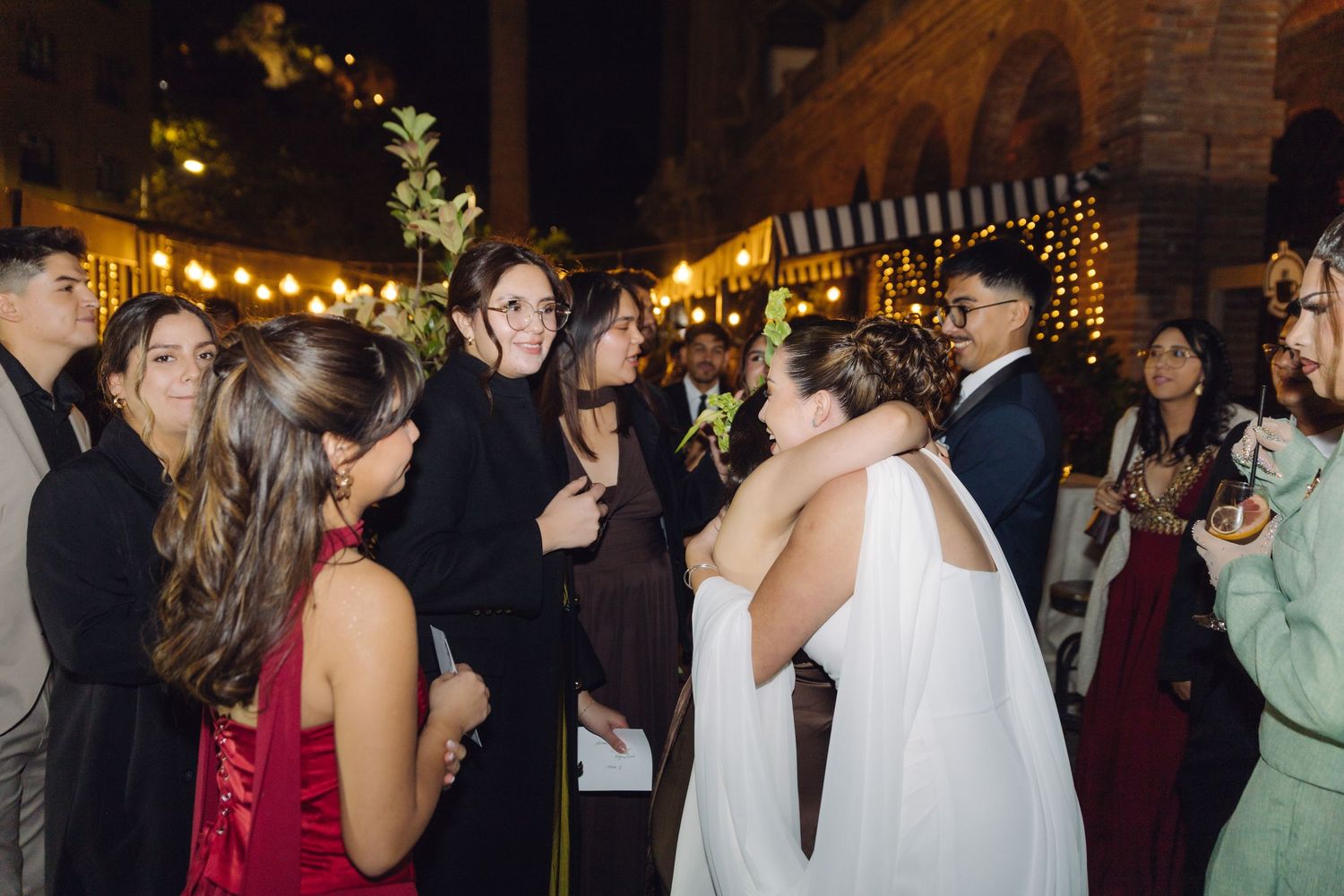 Wedding guests hugging and celebrating with the bride outdoors at a nighttime reception with string lights.