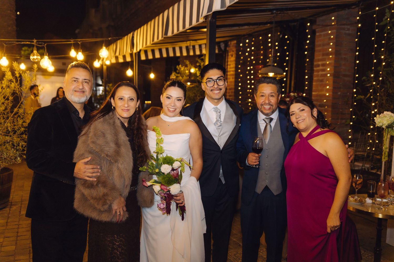 Wedding couple poses with family members outdoors at night, surrounded by warm string lights.