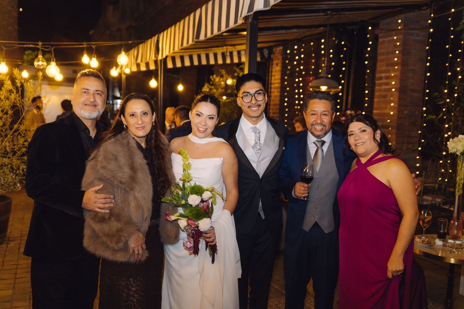 Newlyweds smile with family group at elegant outdoor evening wedding reception venue.