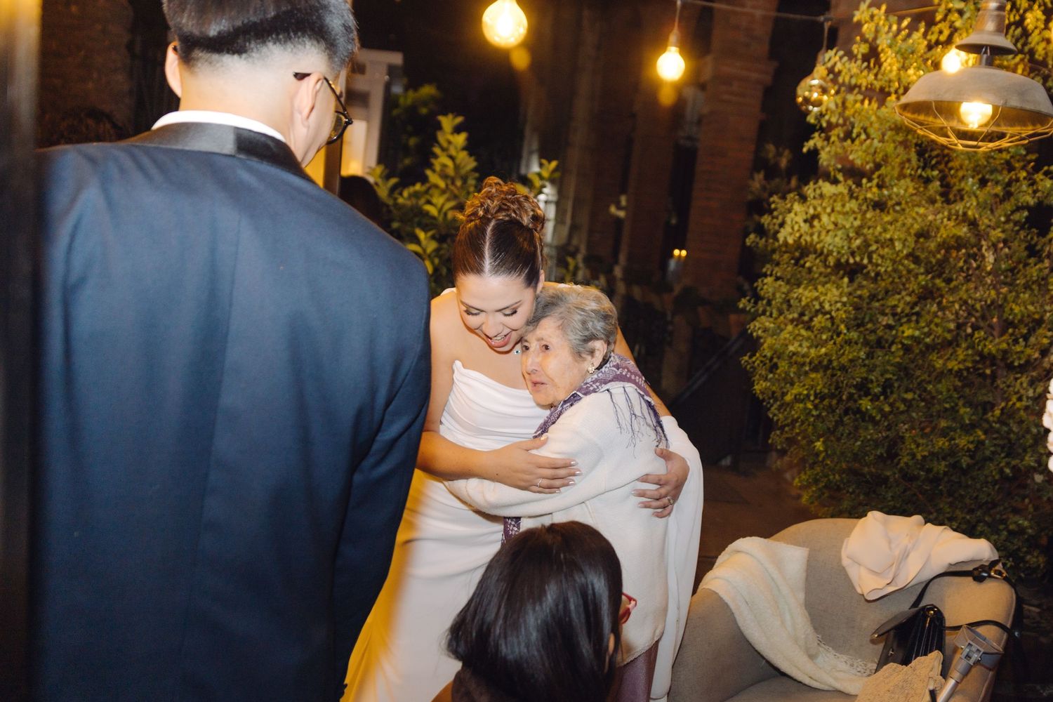 Bride in white gown shares heartfelt hug with elderly guest at outdoor wedding reception.