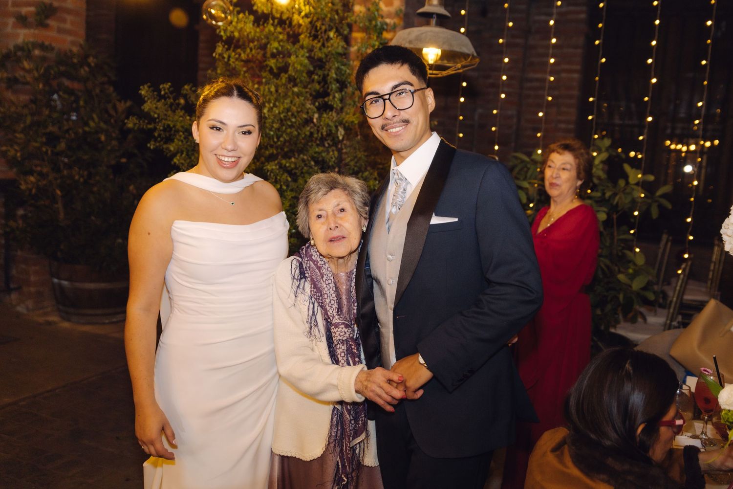 Bride and groom pose with elderly grandmother holding hands at outdoor wedding celebration.