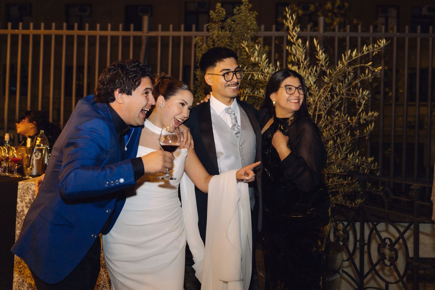 Wedding couple laughs with guests holding wine glasses at outdoor evening reception.