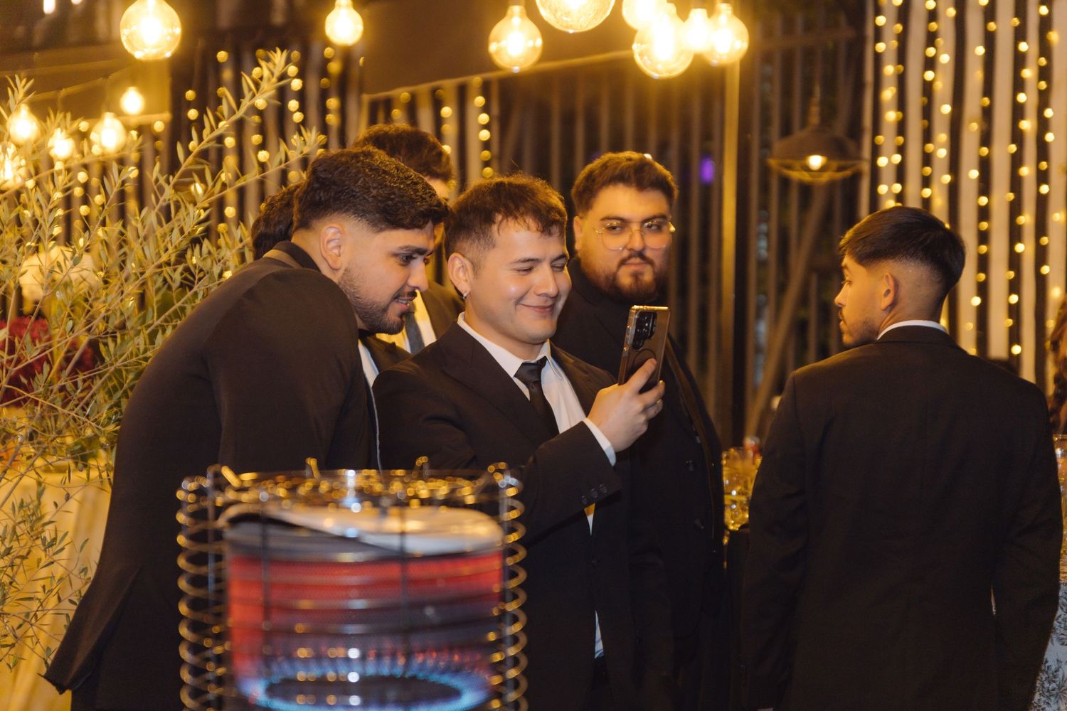 Groomsmen in black suits gather around smartphone at decorated outdoor wedding reception.