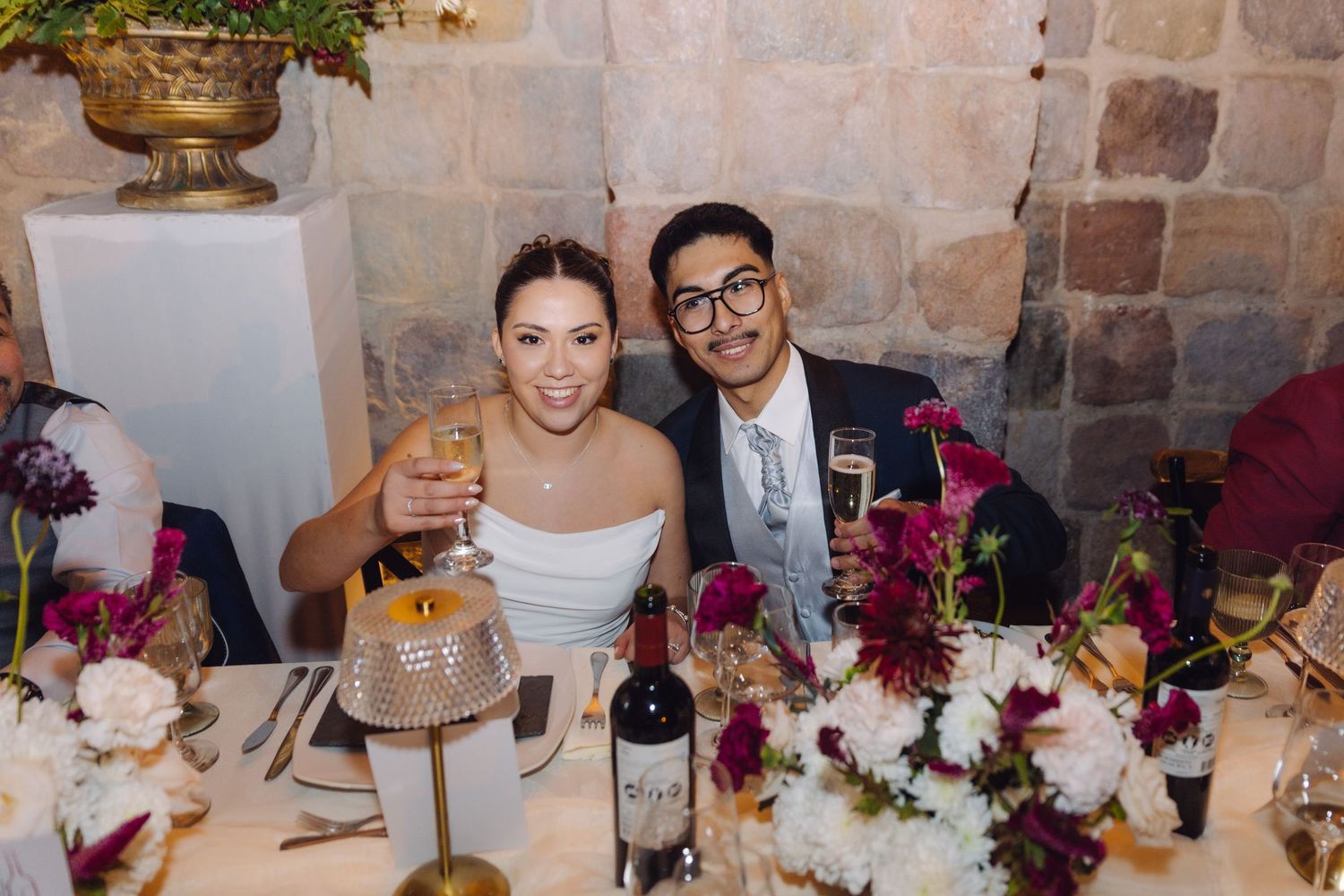Newlyweds toast champagne glasses at floral-decorated head table inside stone venue.