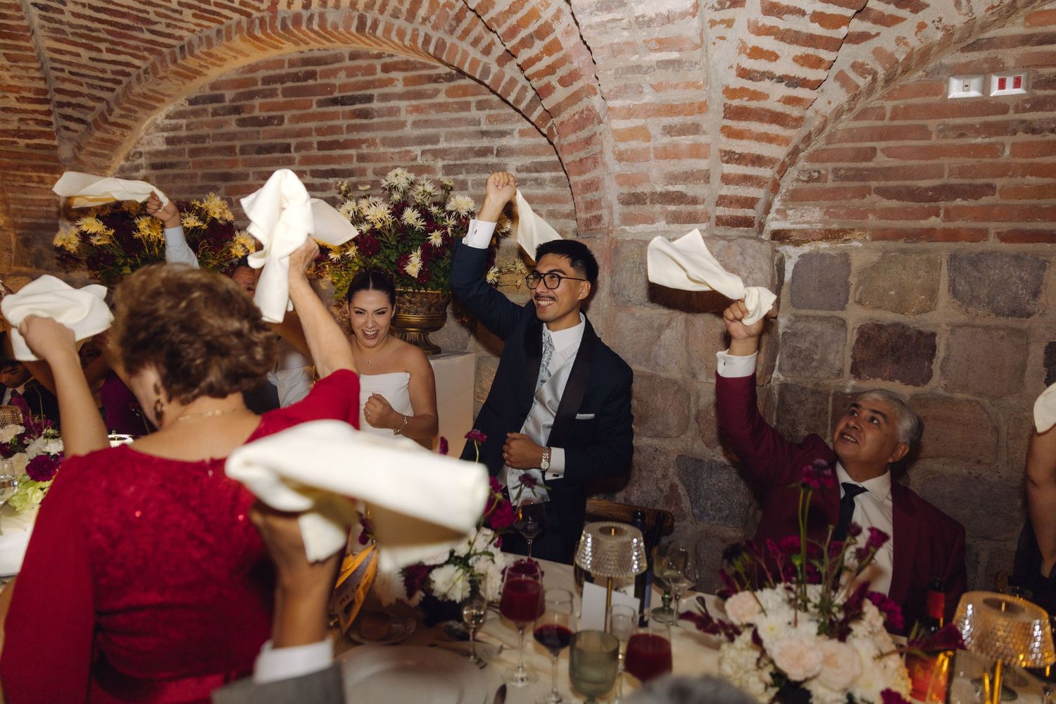 Wedding guests wave white napkins in celebration under brick arched ceiling at reception.
