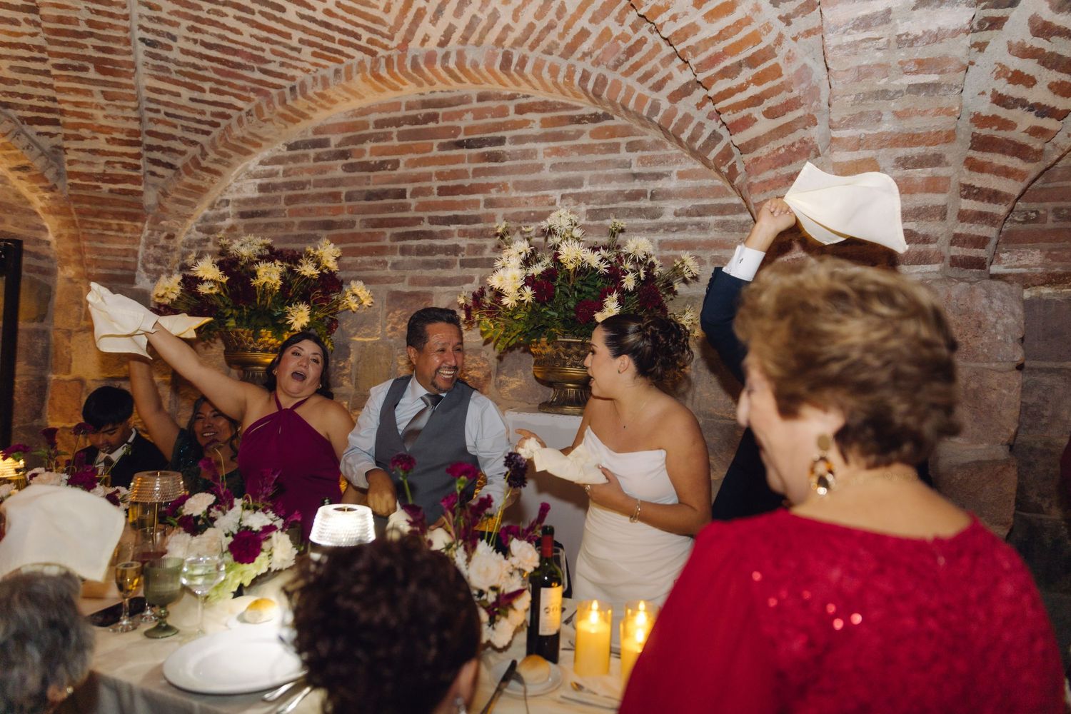 Joyful wedding party waves napkins at head table inside rustic brick arched reception hall.